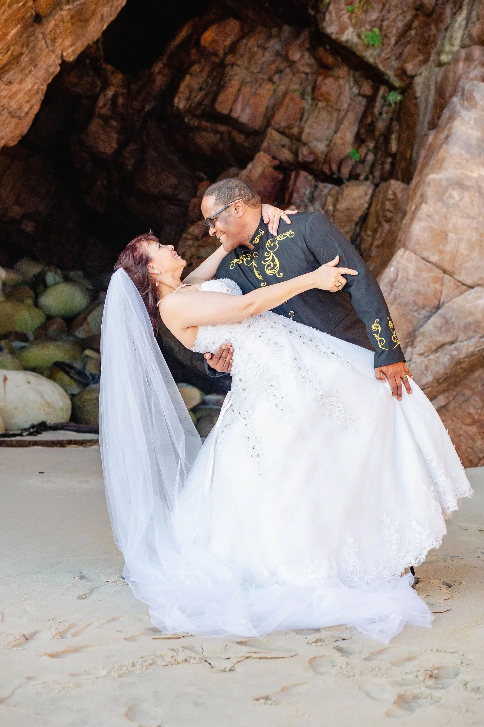 Groom in a black jacket with gold embroidery dipping his bride in the mouth of a sea cave, her veil spread on the sand.