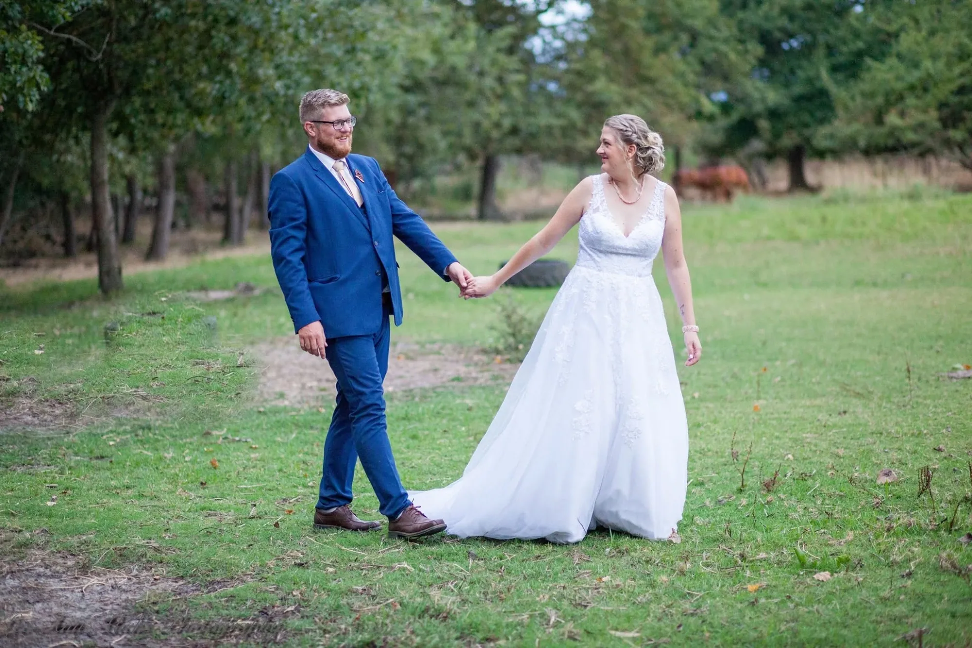 Groom in a blue suit leading his bride by the hand across a green lawn edged with trees.