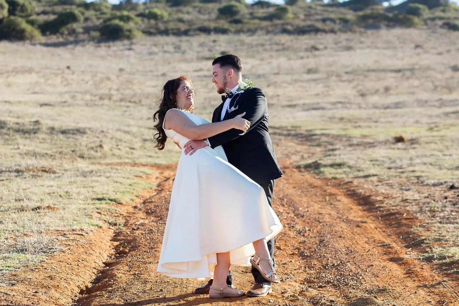 Groom lifting his laughing bride off a red dirt track through dry grass, her tea-length skirt swinging out.