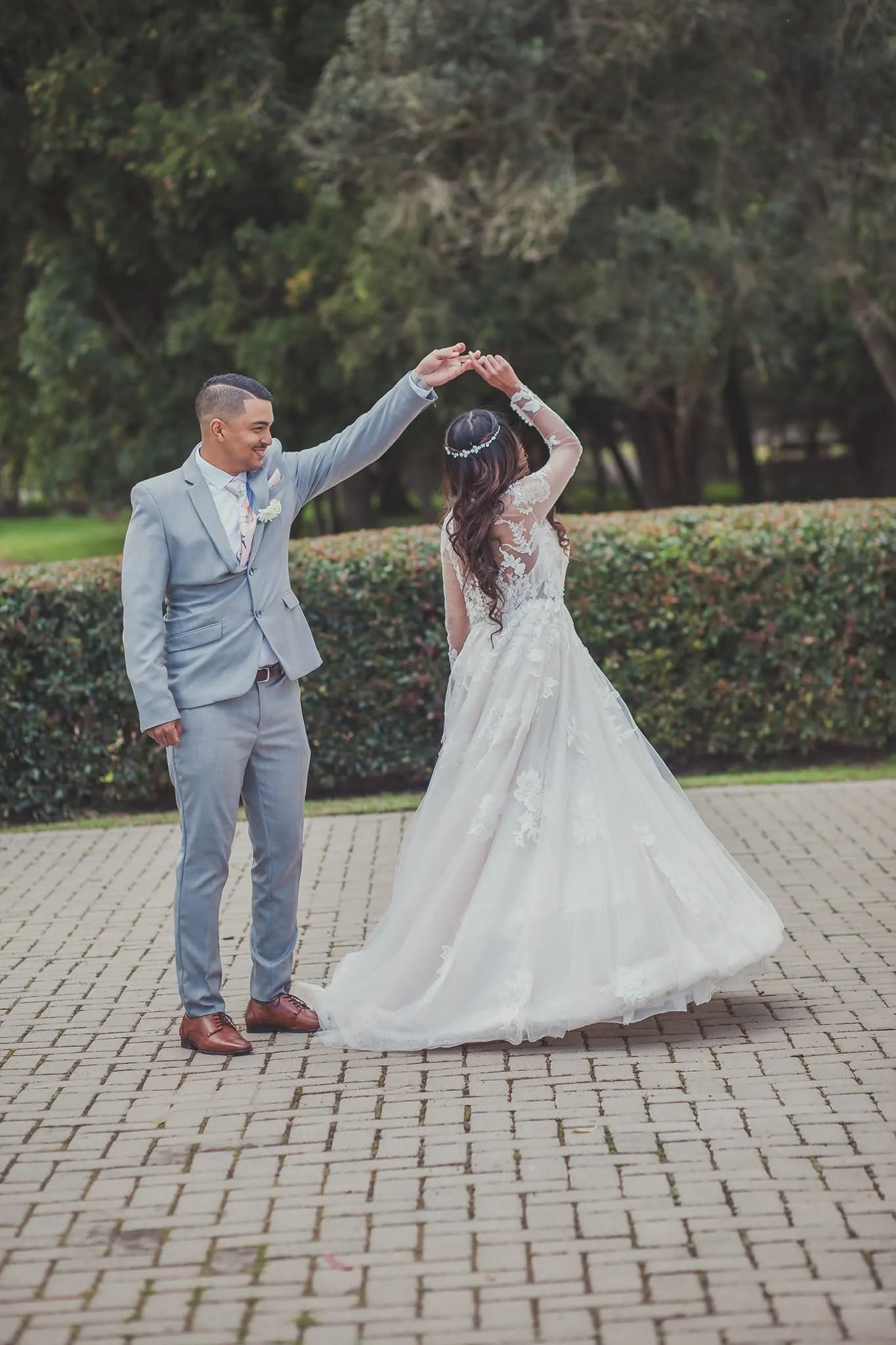 Groom in a pale grey suit twirling his bride on brick paving in front of a clipped hedge.