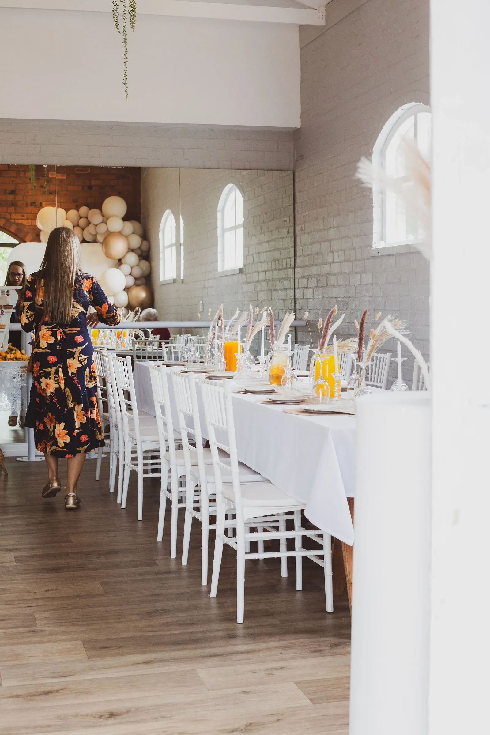 A guest in a floral dress walking beside the long table towards the balloon arch in a bright hall with arched windows.