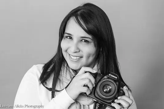 Black and white studio portrait of Lee-Ann Alicks, Cape Town photographer, smiling and holding her Canon camera at chest height against a plain grey backdrop.