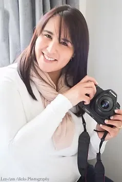 Colour portrait of Lee-Ann Alicks, Somerset West wedding and lifestyle photographer, in a white top and blush scarf, holding her camera beside a grey curtain.