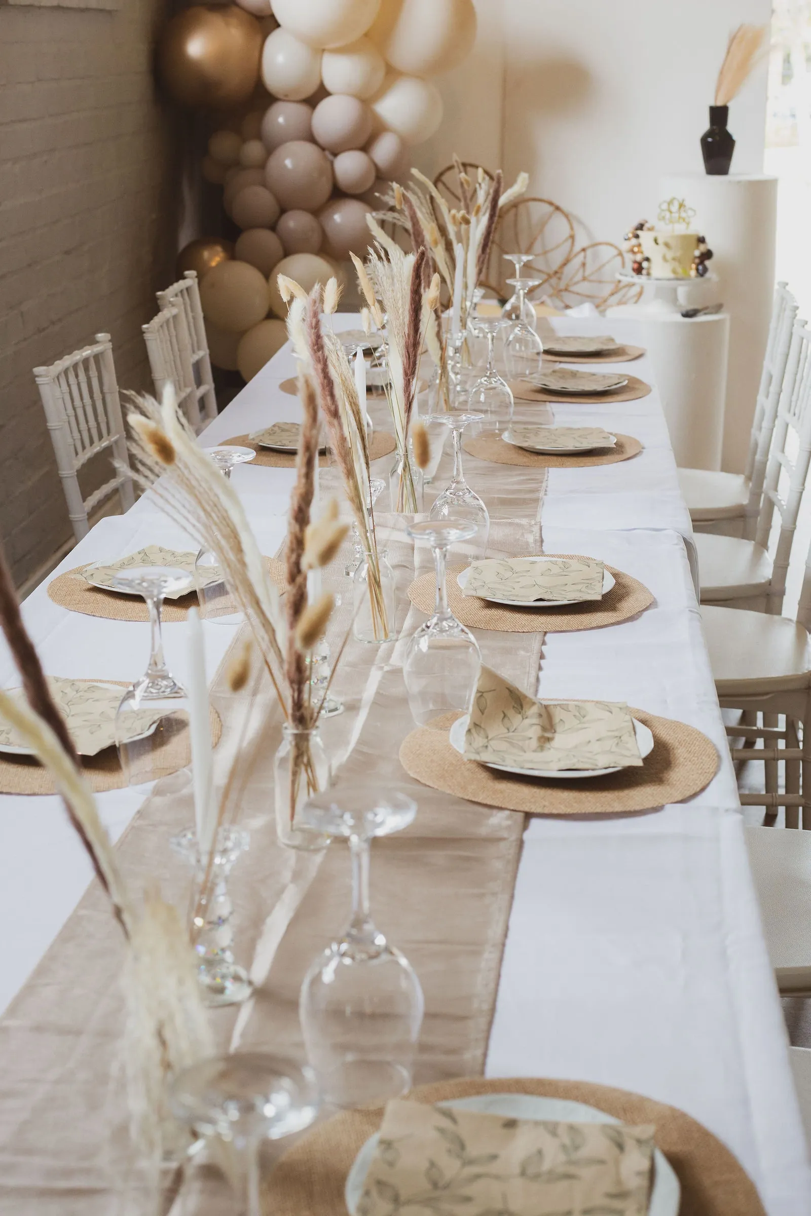 View down a styled long table with jute place mats, leaf-print napkins and pampas stems leading to a balloon garland and a cake on a plinth.