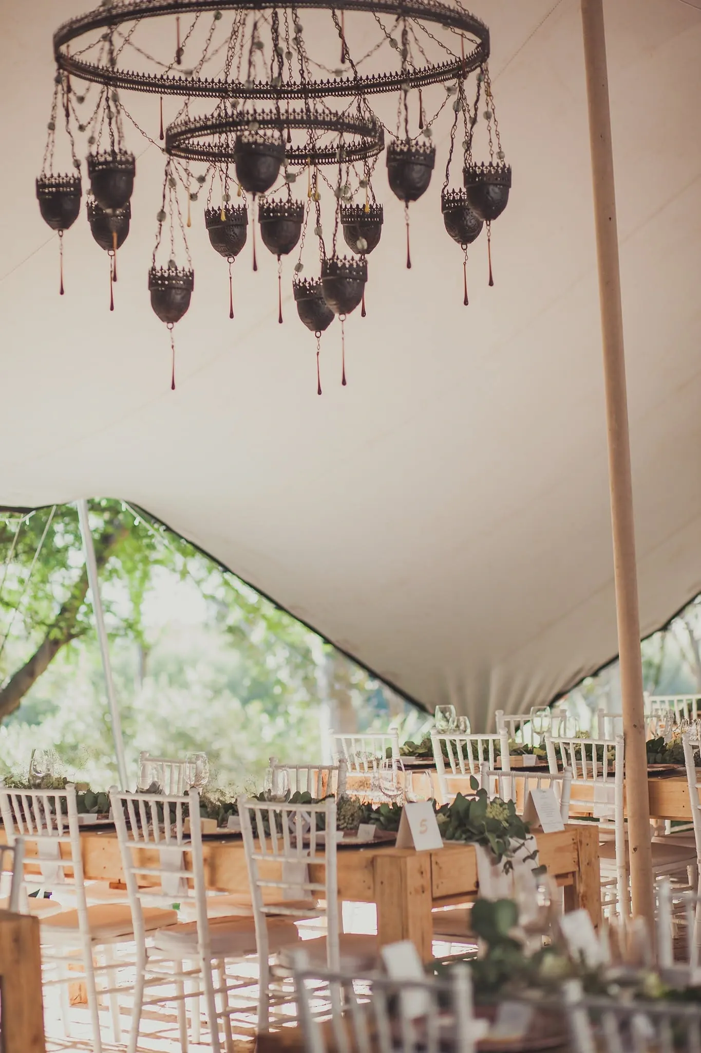 Stretch-tent reception with a wrought-iron chandelier above long wooden tables and white chairs.