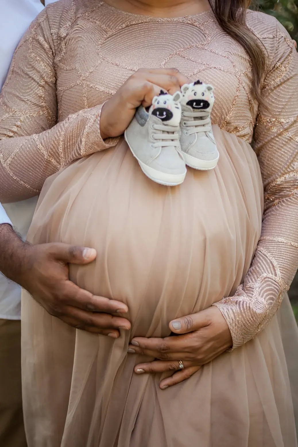 Pair of tiny baby sneakers held against a blush tulle maternity gown, a partner's hand on the bump.