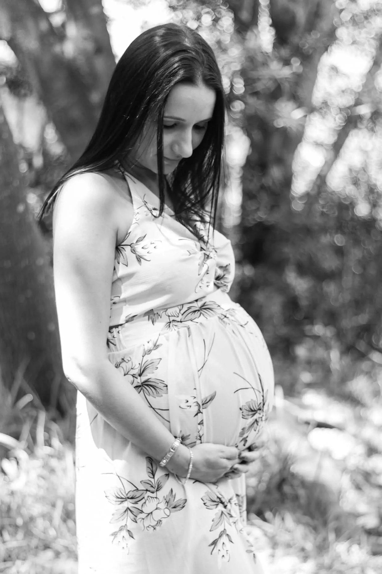 Black and white photo of a mum-to-be in a floral halter dress looking down at her bump under trees.