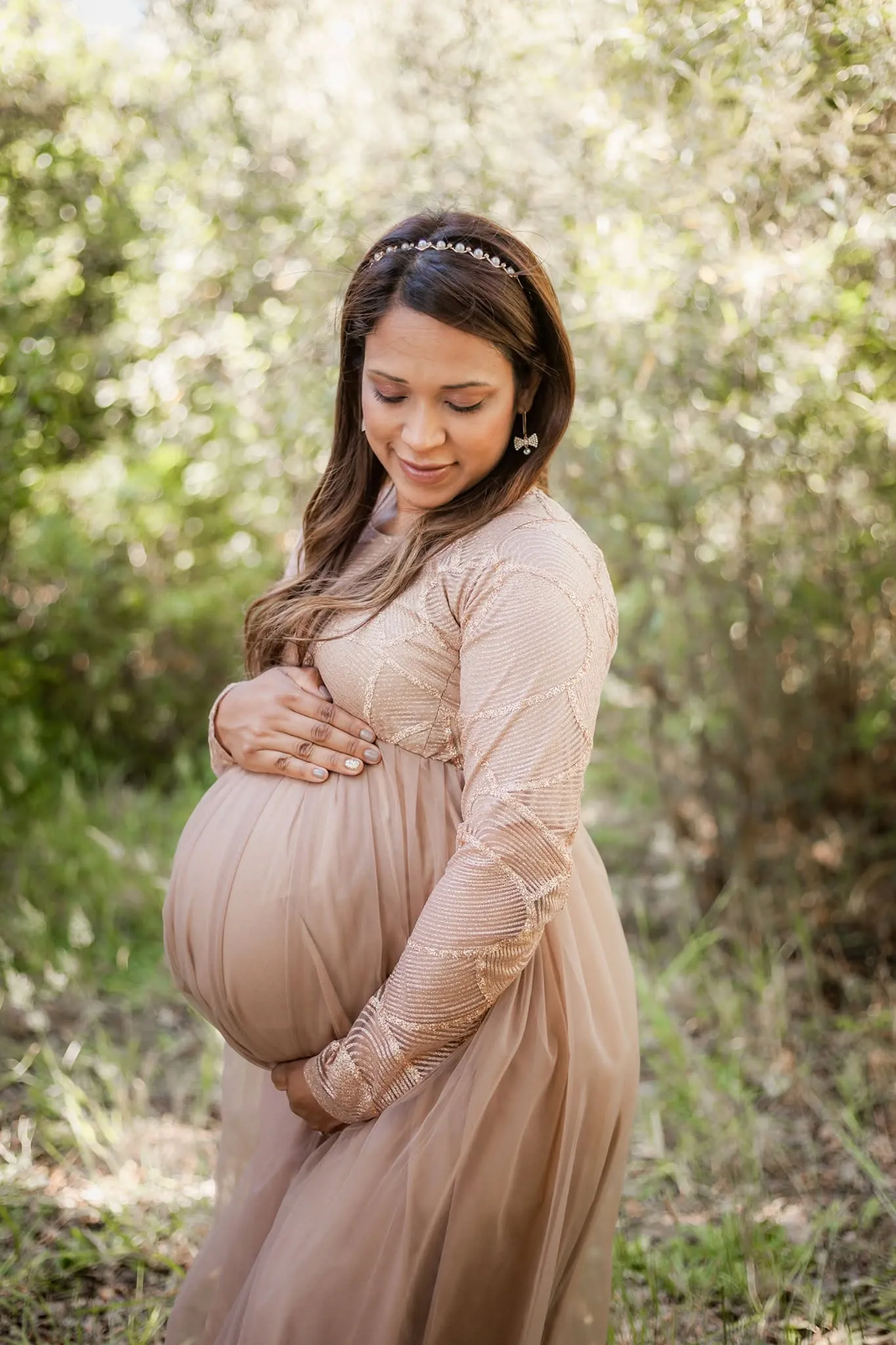 Mum-to-be in a blush tulle gown and jewelled headband looking down at her bump in a sunny woodland clearing.