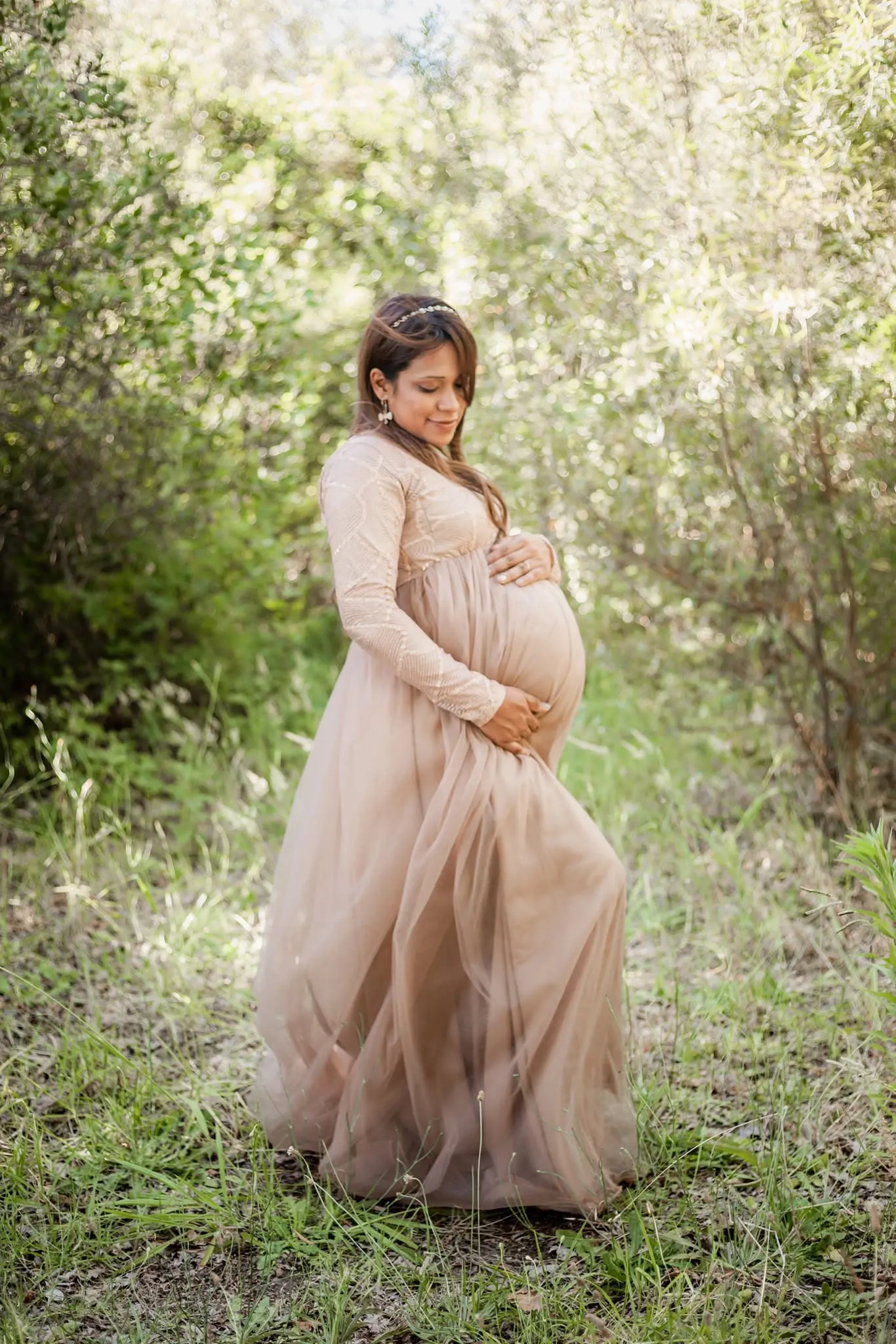 Mum-to-be in a blush tulle gown with lace sleeves, hands on her bump, in long grass.