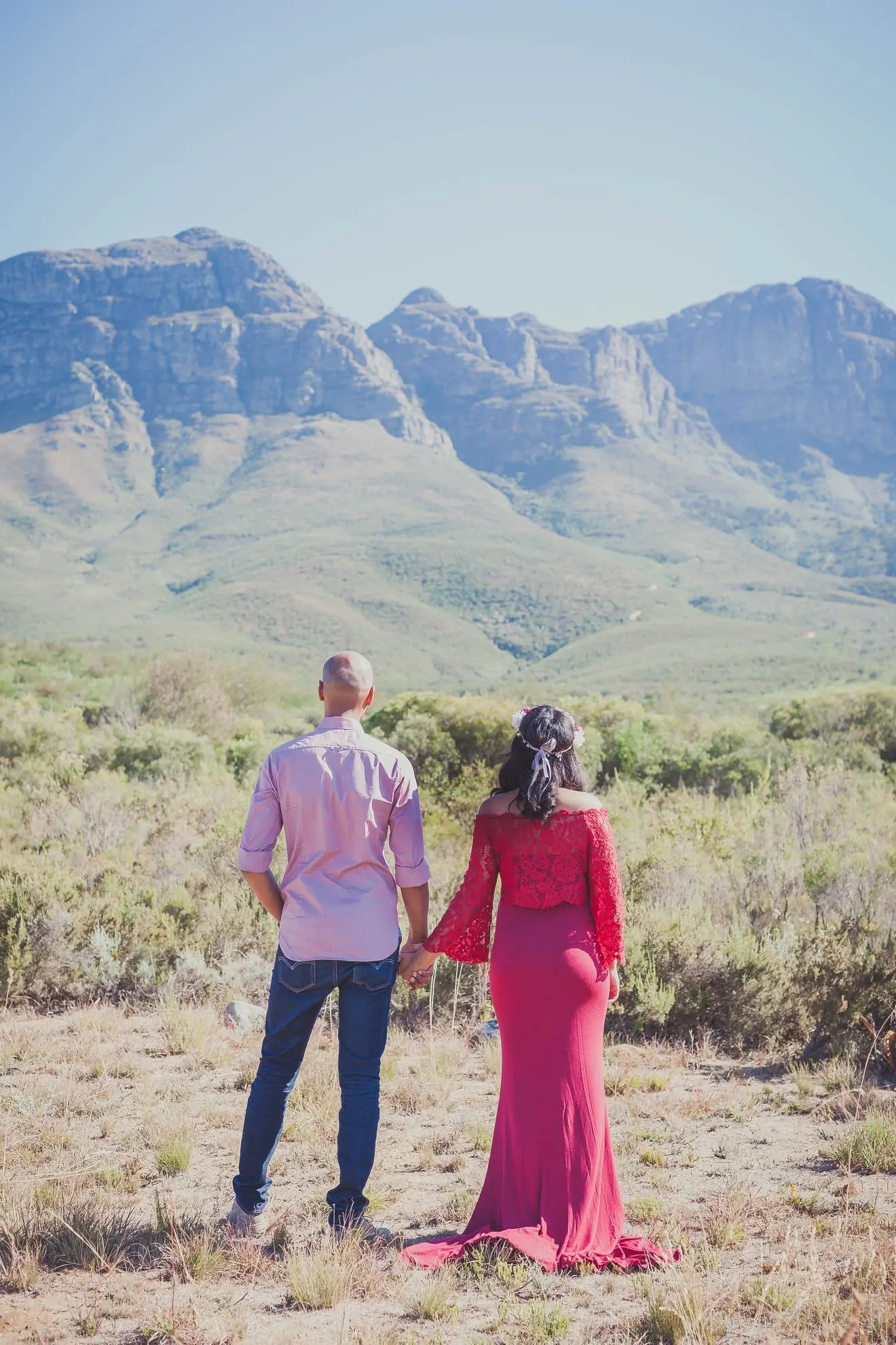 Expecting couple seen from behind, holding hands and facing a range of rocky mountains, her red gown trailing on the ground.