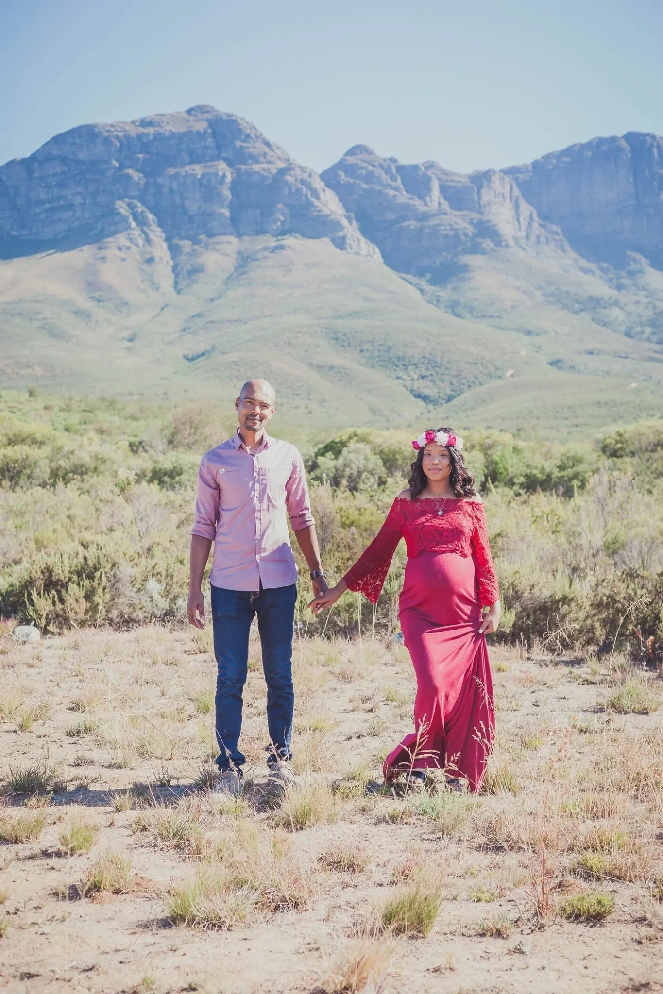 Expecting couple holding hands in dry veld, she in a red lace maternity gown and flower crown, rocky peaks behind.