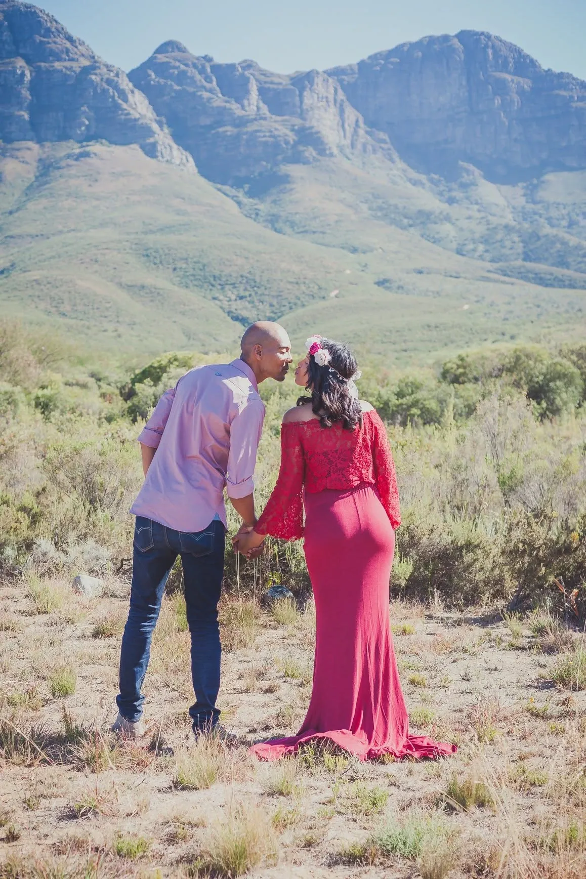 Expecting couple kissing in dry veld, her red gown pooling behind her, with tall mountains in the distance.