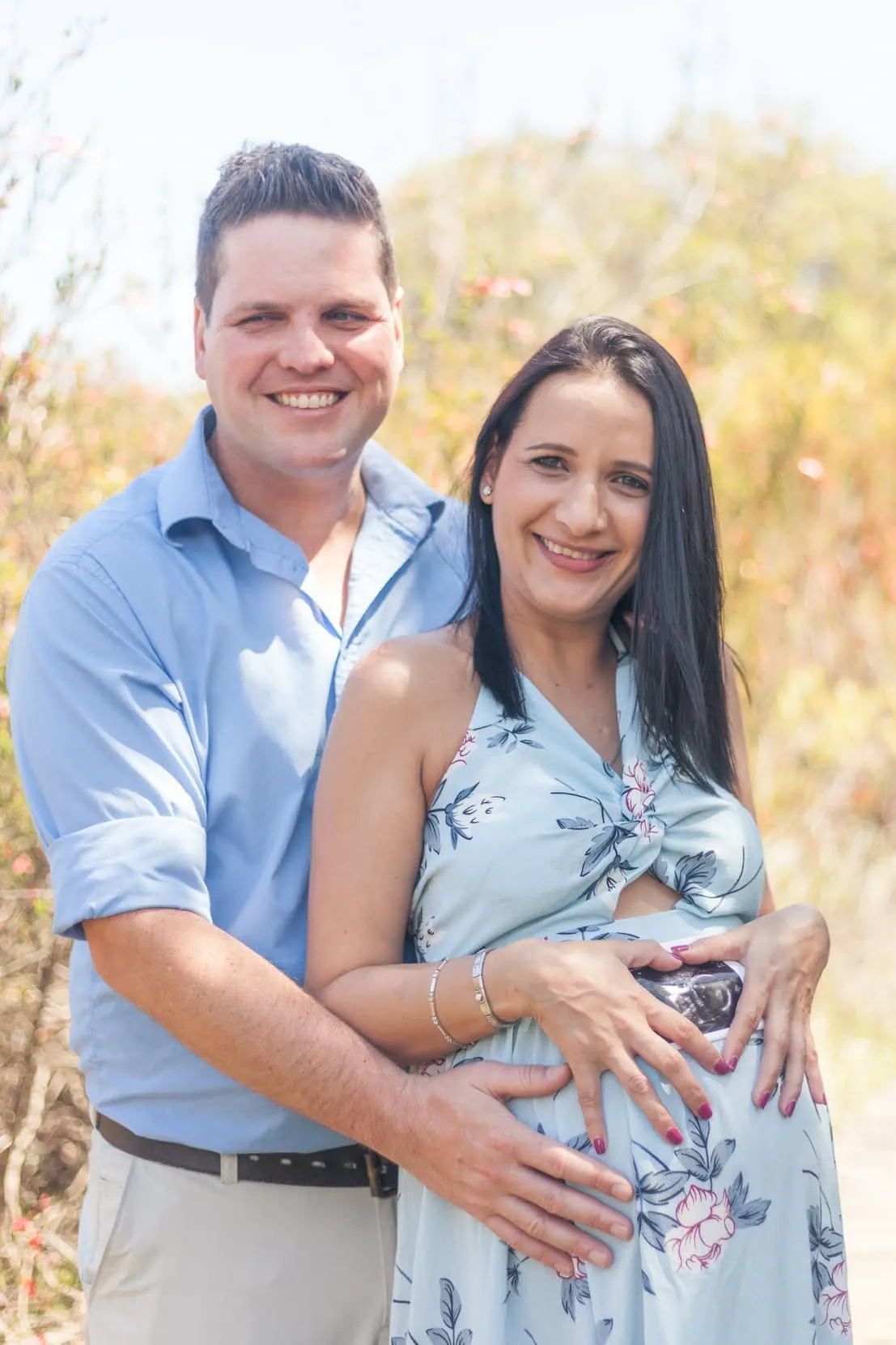 Smiling expecting couple, her hands holding a scan print against the bump and his hand beneath it.