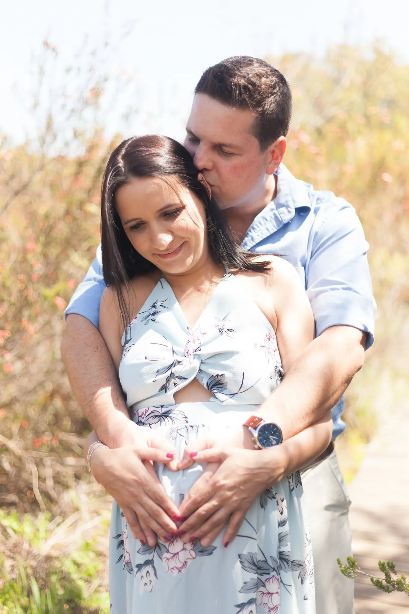 Partner kissing his pregnant wife's temple from behind as they shape a heart with their hands on her bump.