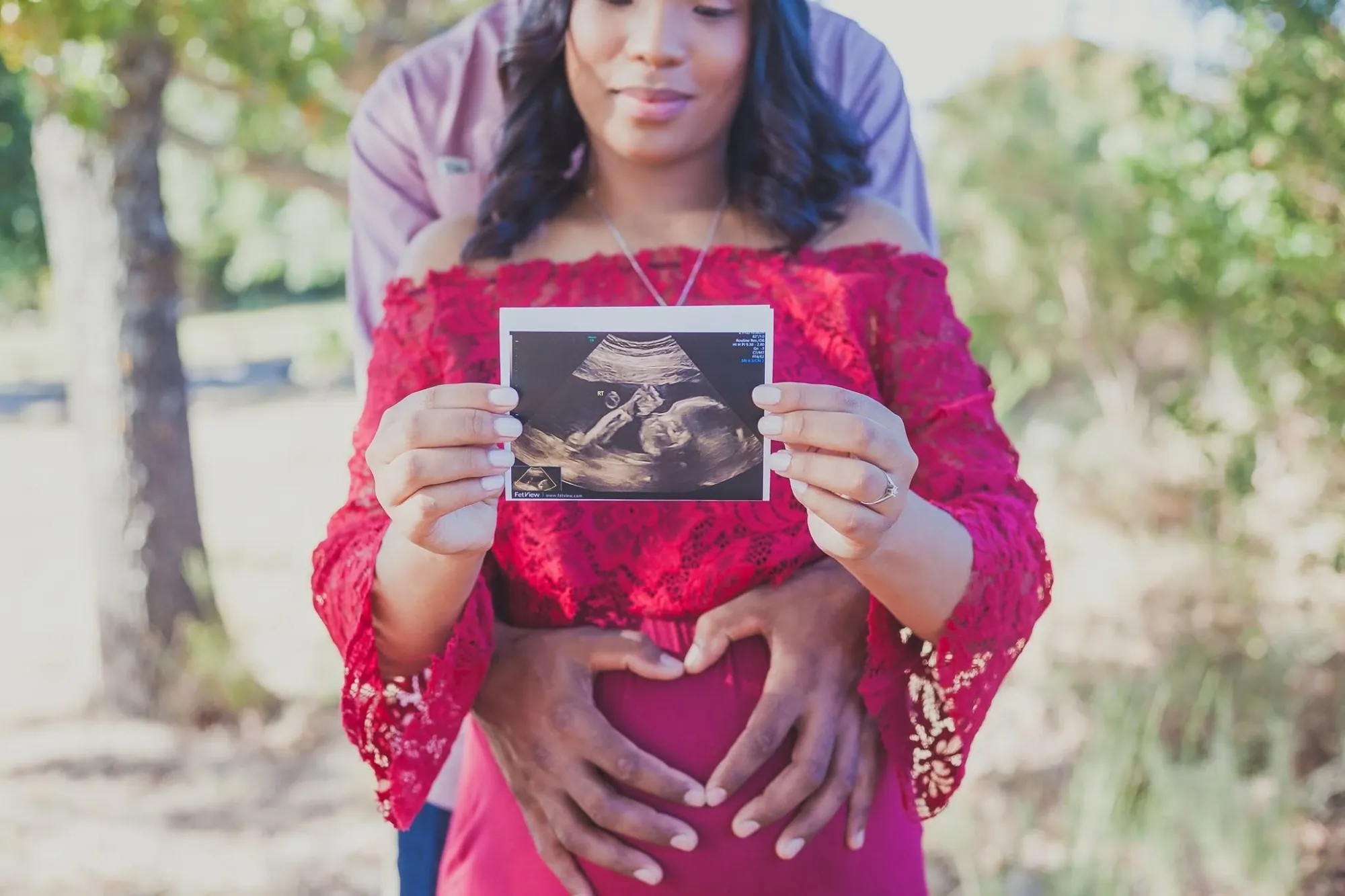 Mum-to-be in a red lace top holding up her baby's scan photo while her partner's hands form a heart on the bump.