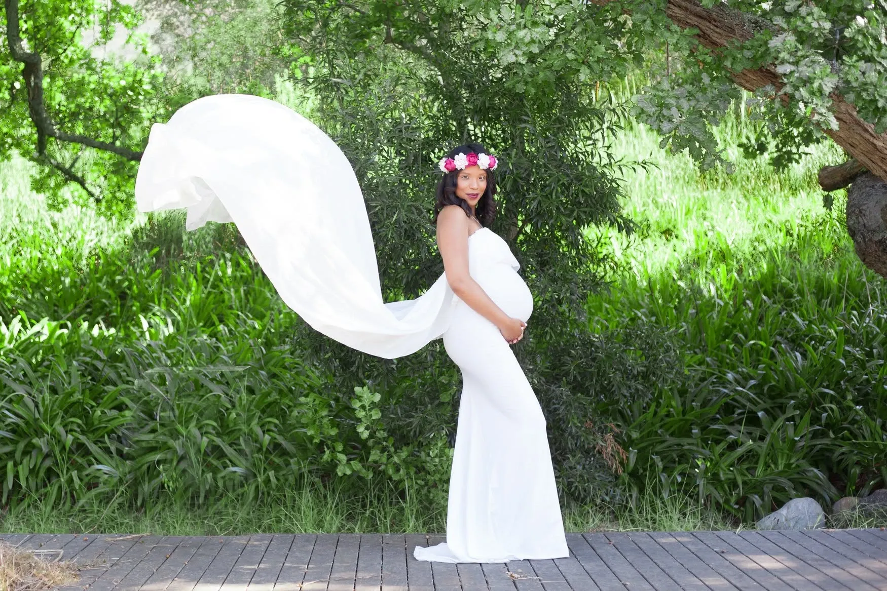 Mum-to-be in a fitted white gown and flower crown, its long sheer train lifted by the wind in a green garden.