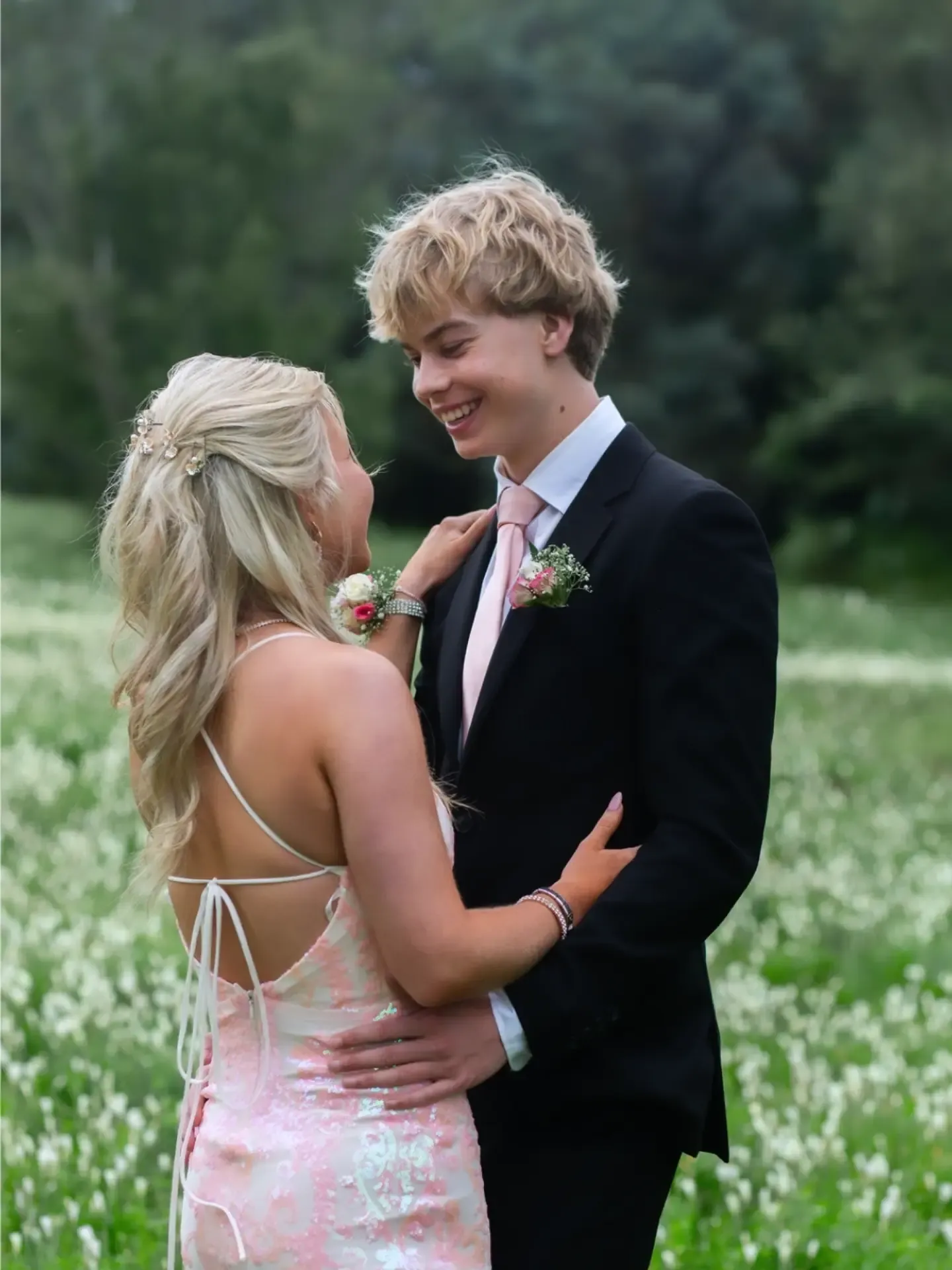 Matric dance couple in a pink sequin gown and black suit smiling at each other in a field of flowers.