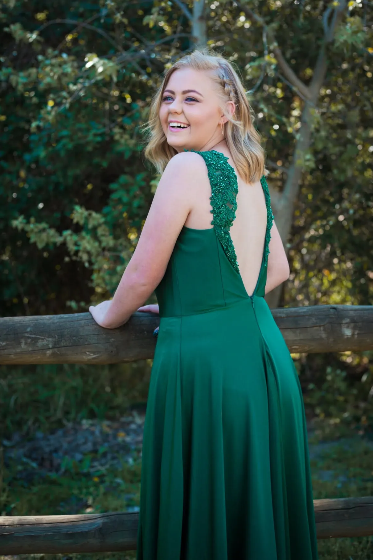 Matriculant in an open-back emerald gown laughing over her shoulder as she leans on a log fence.