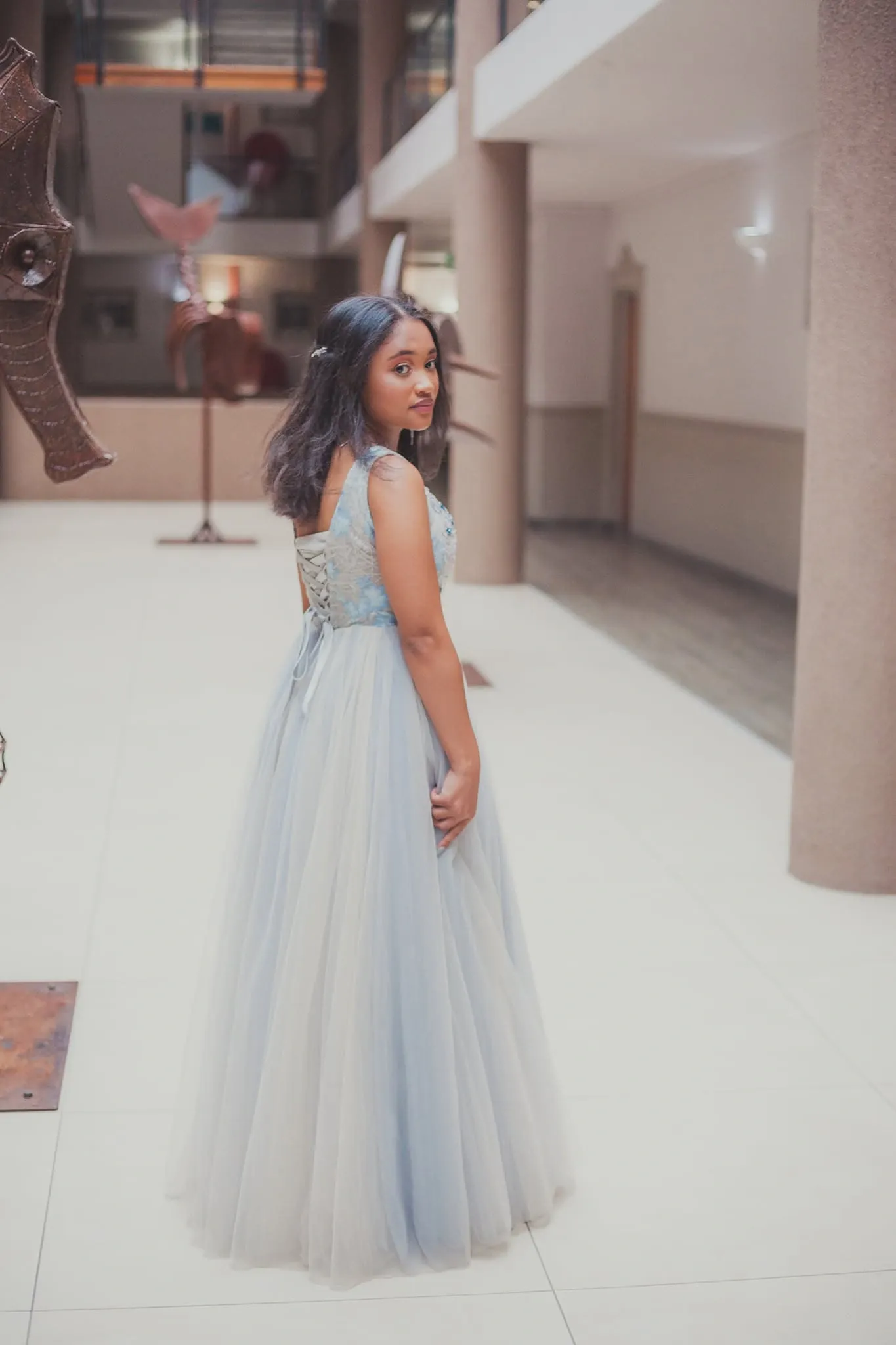 Matriculant in a pale blue tulle gown looking back over her shoulder to show the lace-up back, in a tiled atrium.