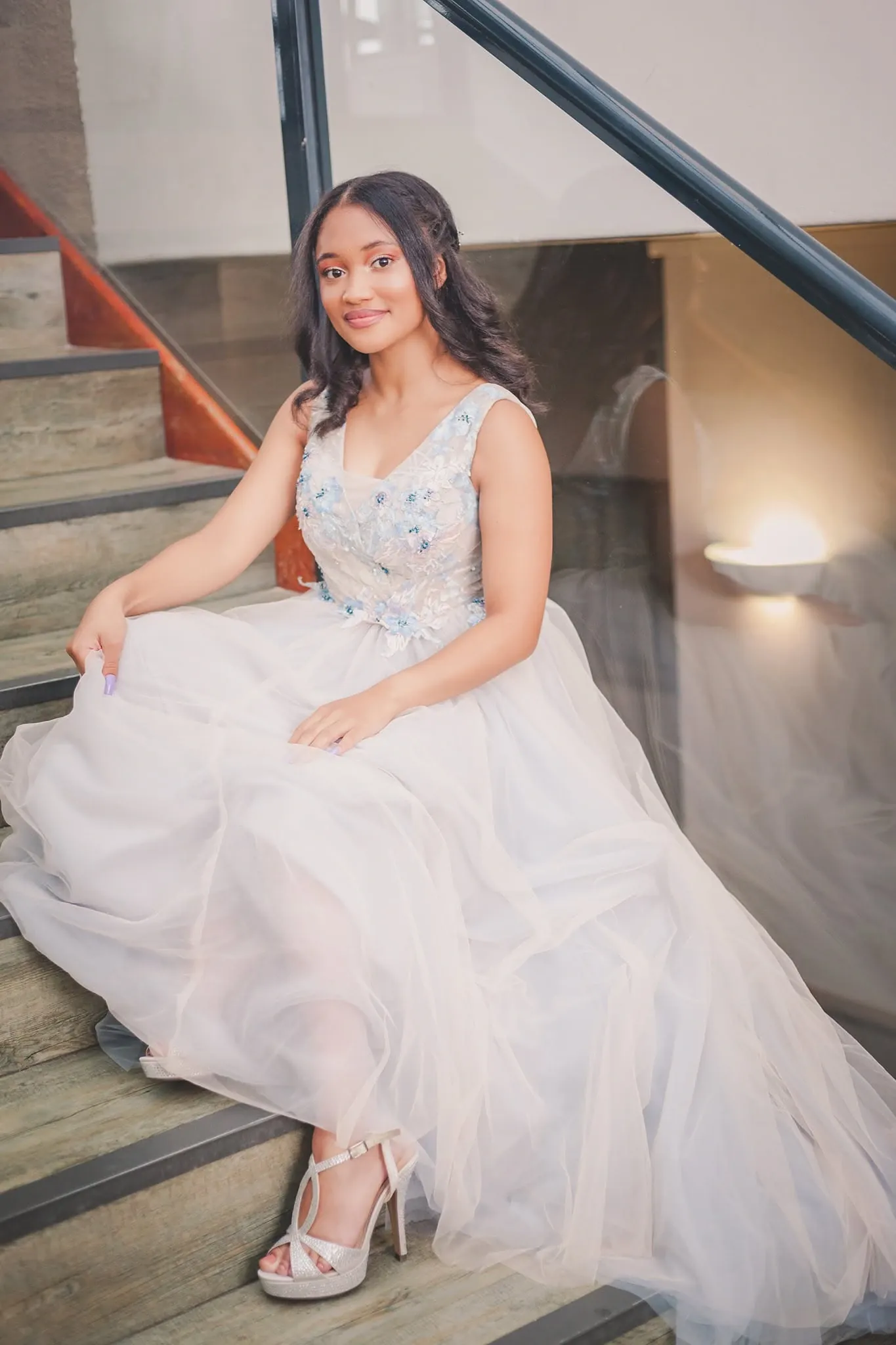 Matriculant in a pale blue tulle gown seated on a wooden staircase, smiling at the camera.