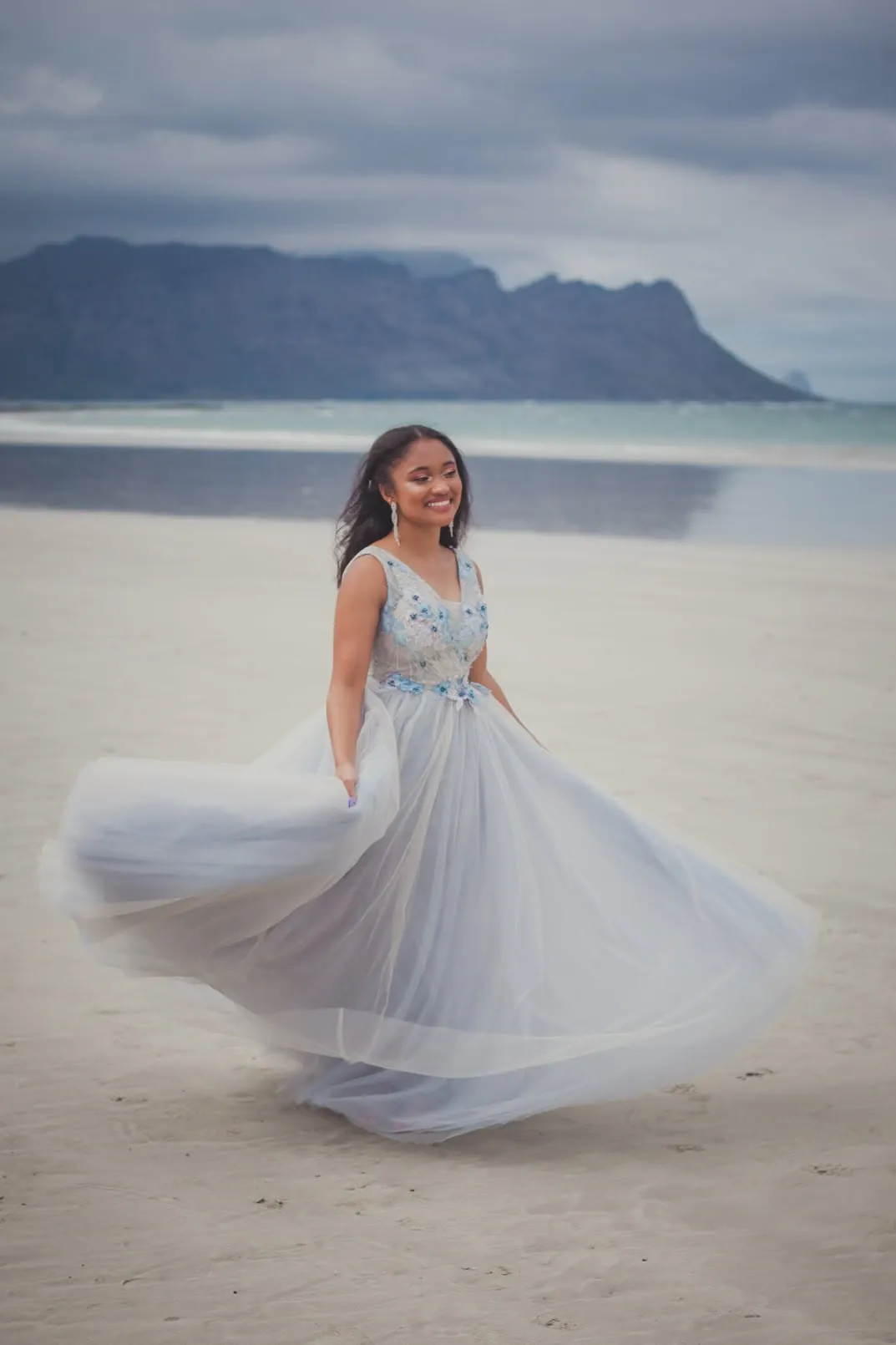 Matriculant twirling her pale blue gown on the beach, mountains across the water behind.