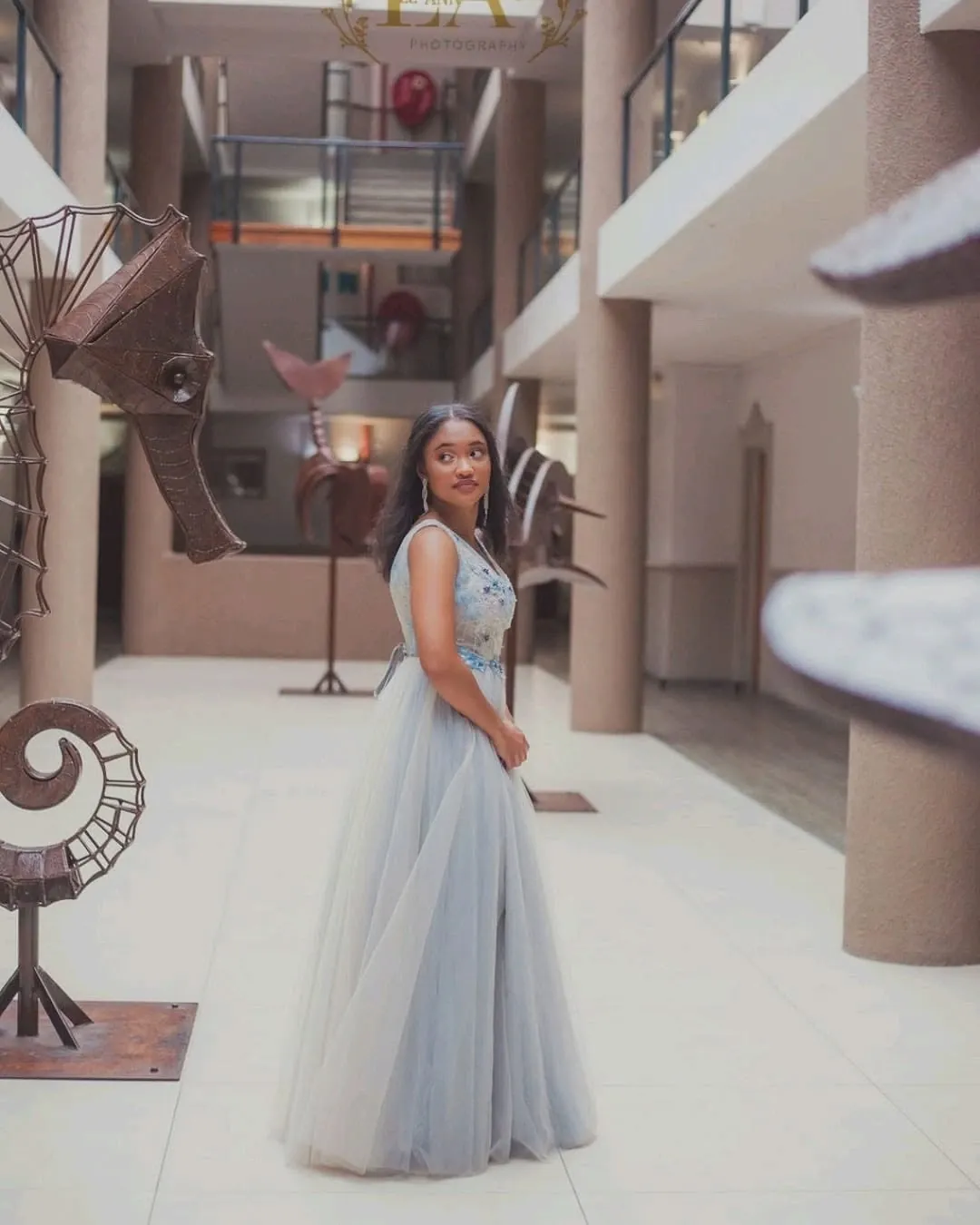 Matriculant in a grey tulle gown looking back over her shoulder in an atrium with metal seahorse sculptures.