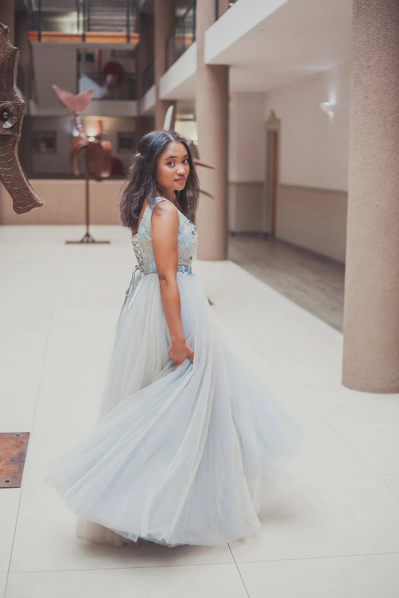 Matriculant twirling her pale blue tulle skirt in a tiled atrium beside a metal seahorse sculpture.