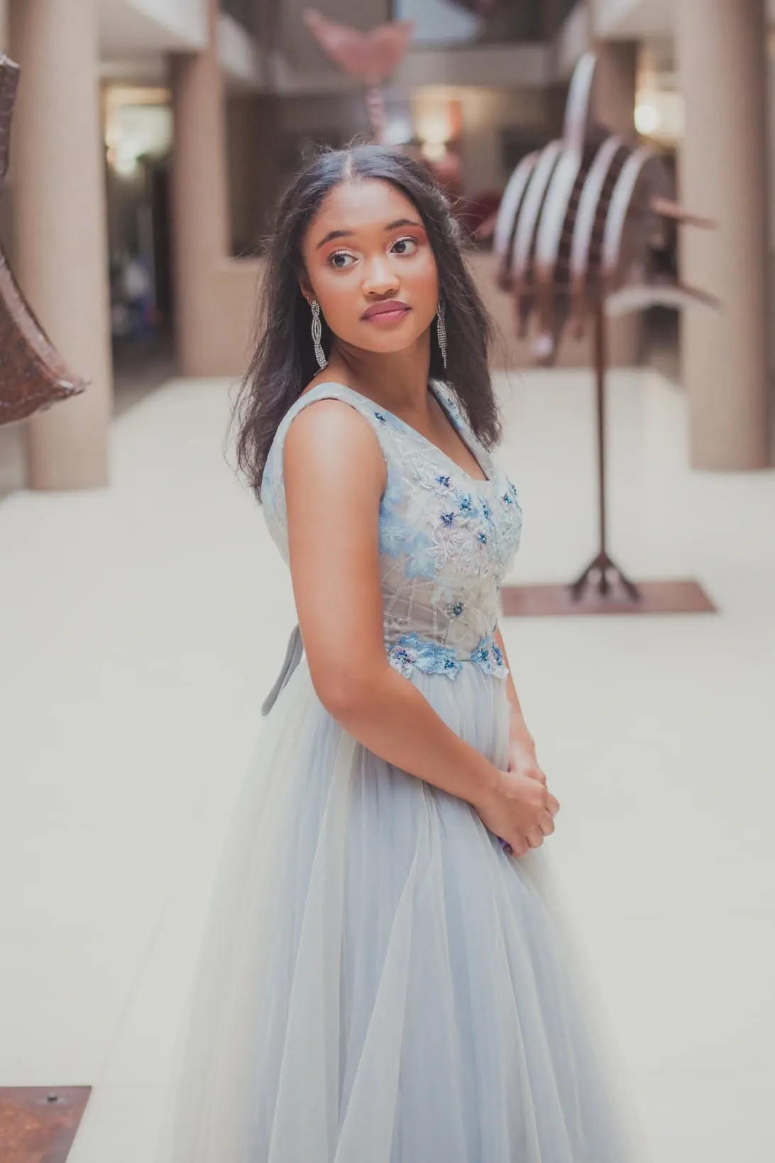 Matriculant in a pale blue gown with floral beading glancing to the side in a tiled atrium with metal sculptures.
