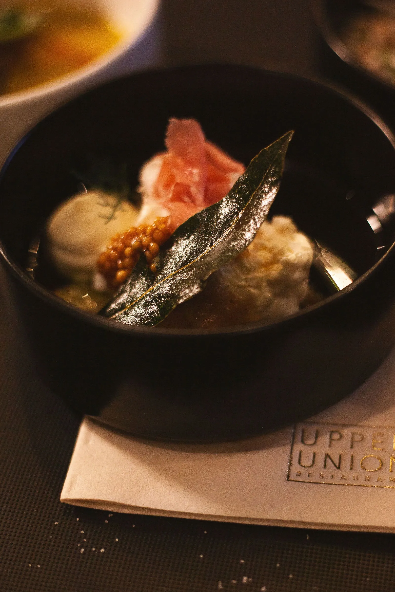 Menu photography: a black bowl holding a folded pink slice, a smooth quenelle, small golden spheres and a glossy dark leaf, a printed napkin beneath.