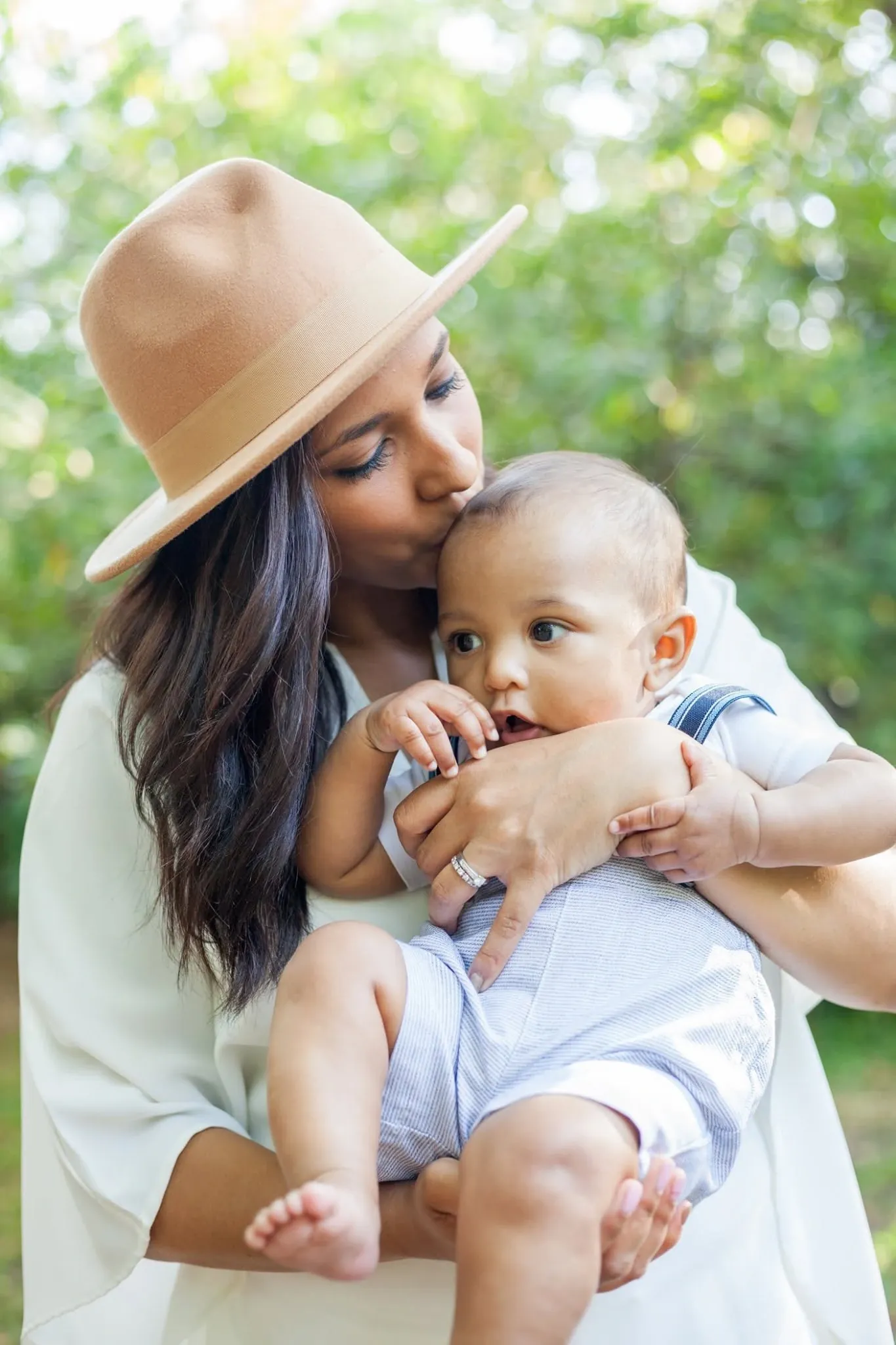 Mother in a tan fedora kissing the top of her baby son's head while he chews on his hand.