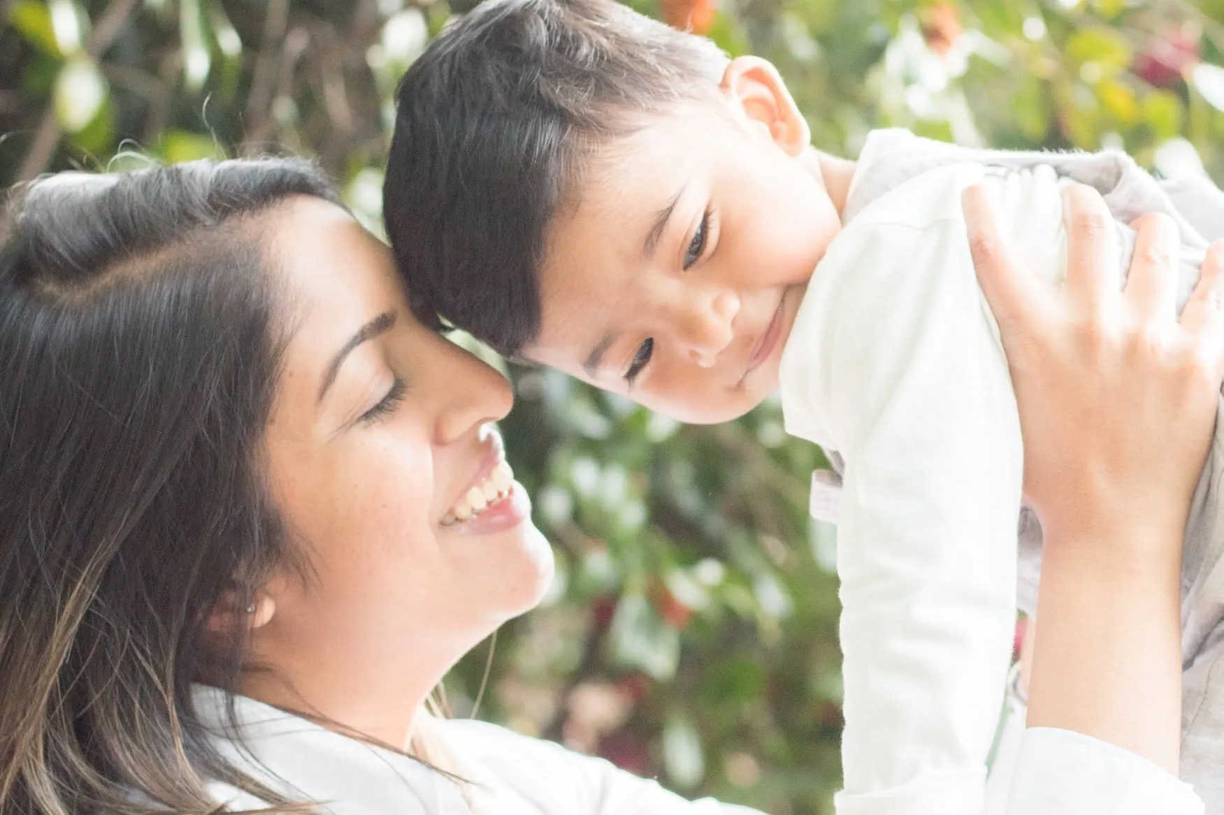 Mother with her eyes closed touching noses with her smiling toddler son as she lifts him, green leaves blurred behind.
