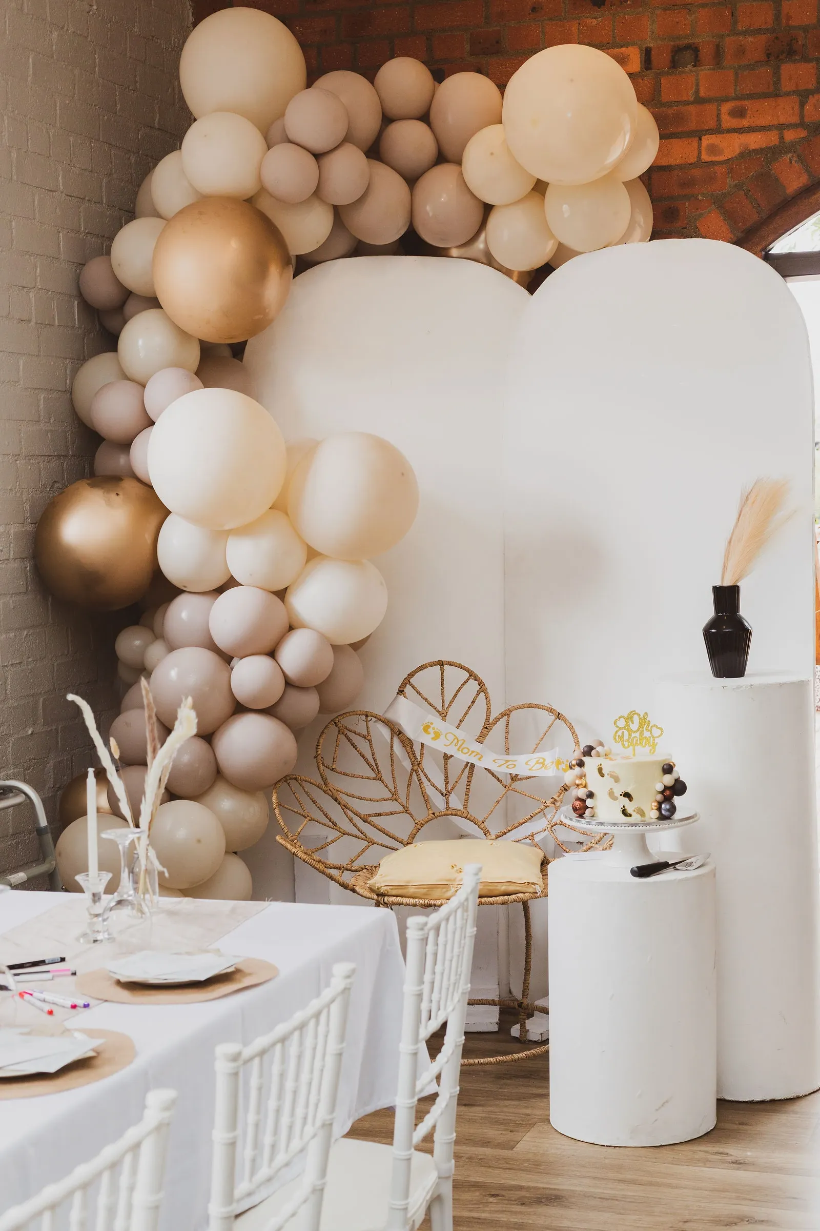 Rattan chair with a Mom To Be sash under a balloon arch against red brick, the cake on a white plinth beside it.
