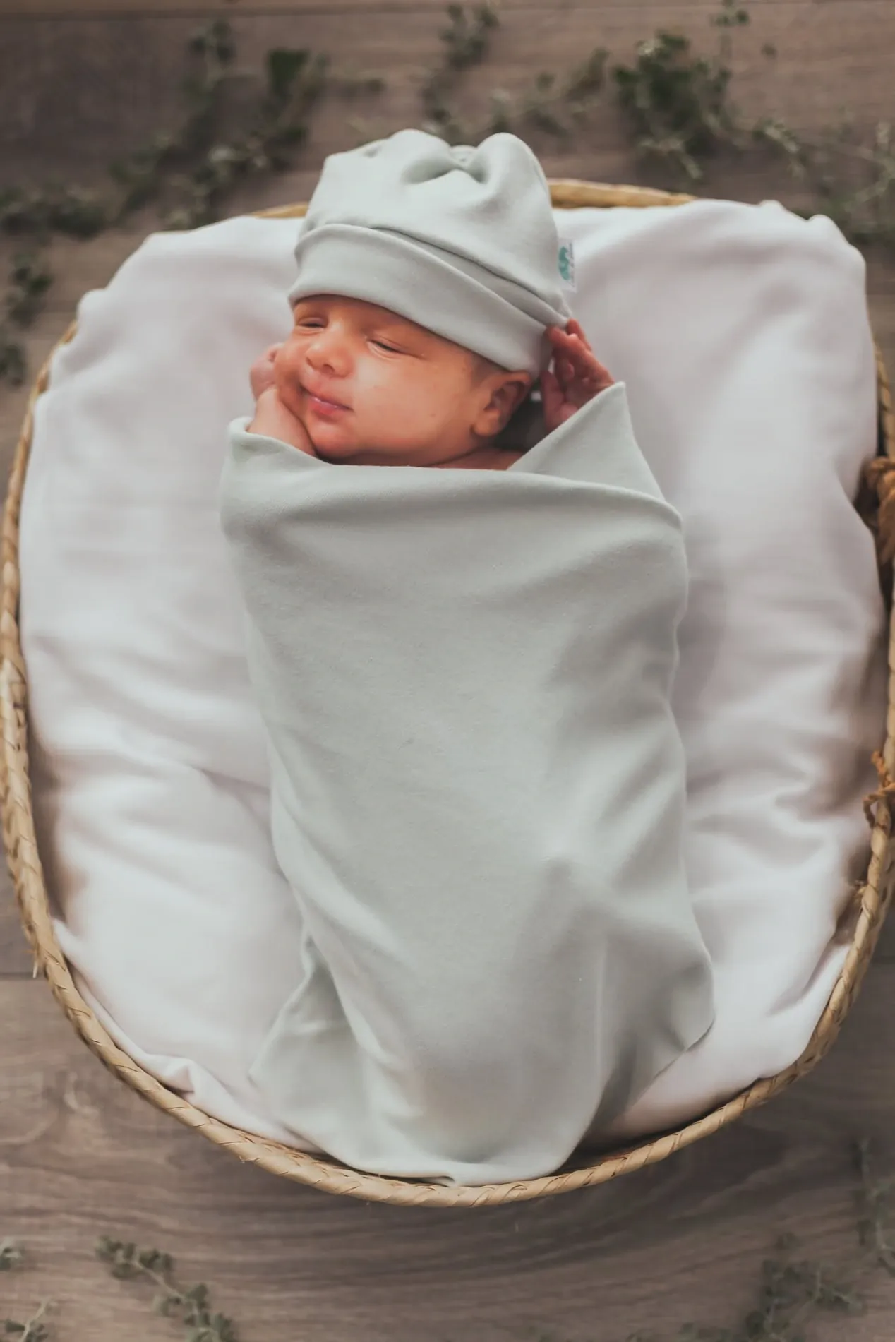 Newborn asleep in a woven basket, swaddled in a soft mint wrap and matching hat.