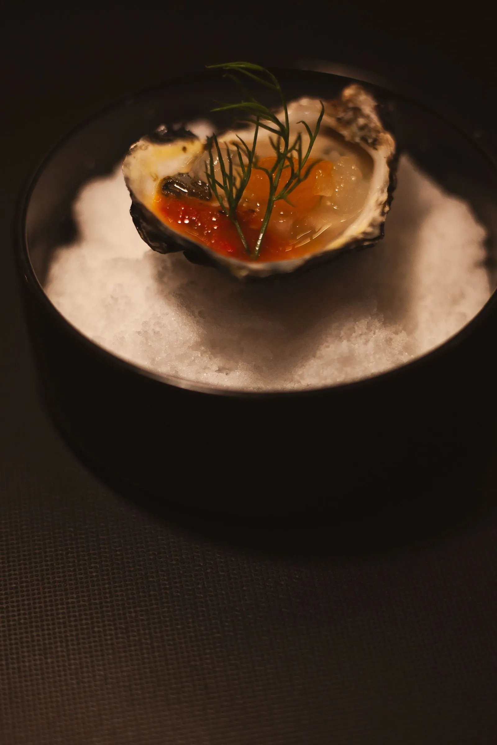 Restaurant photography: an oyster on a frosty white bed in a matte black bowl, topped with amber pearls and a feathery green sprig.