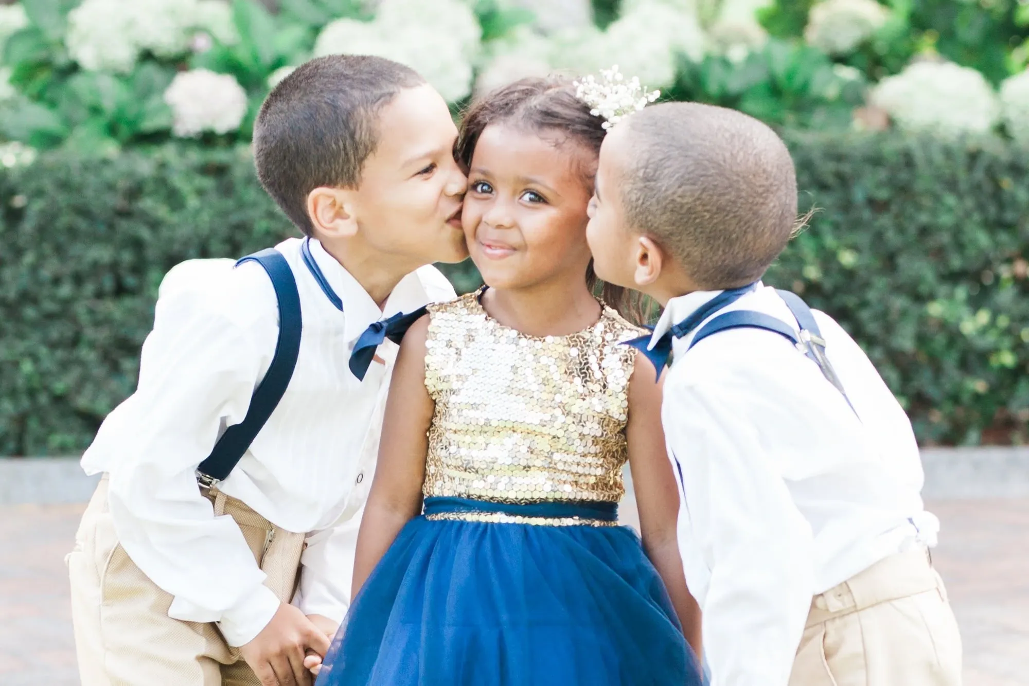 Two pageboys in braces and bow ties kissing the cheeks of a smiling flower girl in a gold sequin and navy dress.