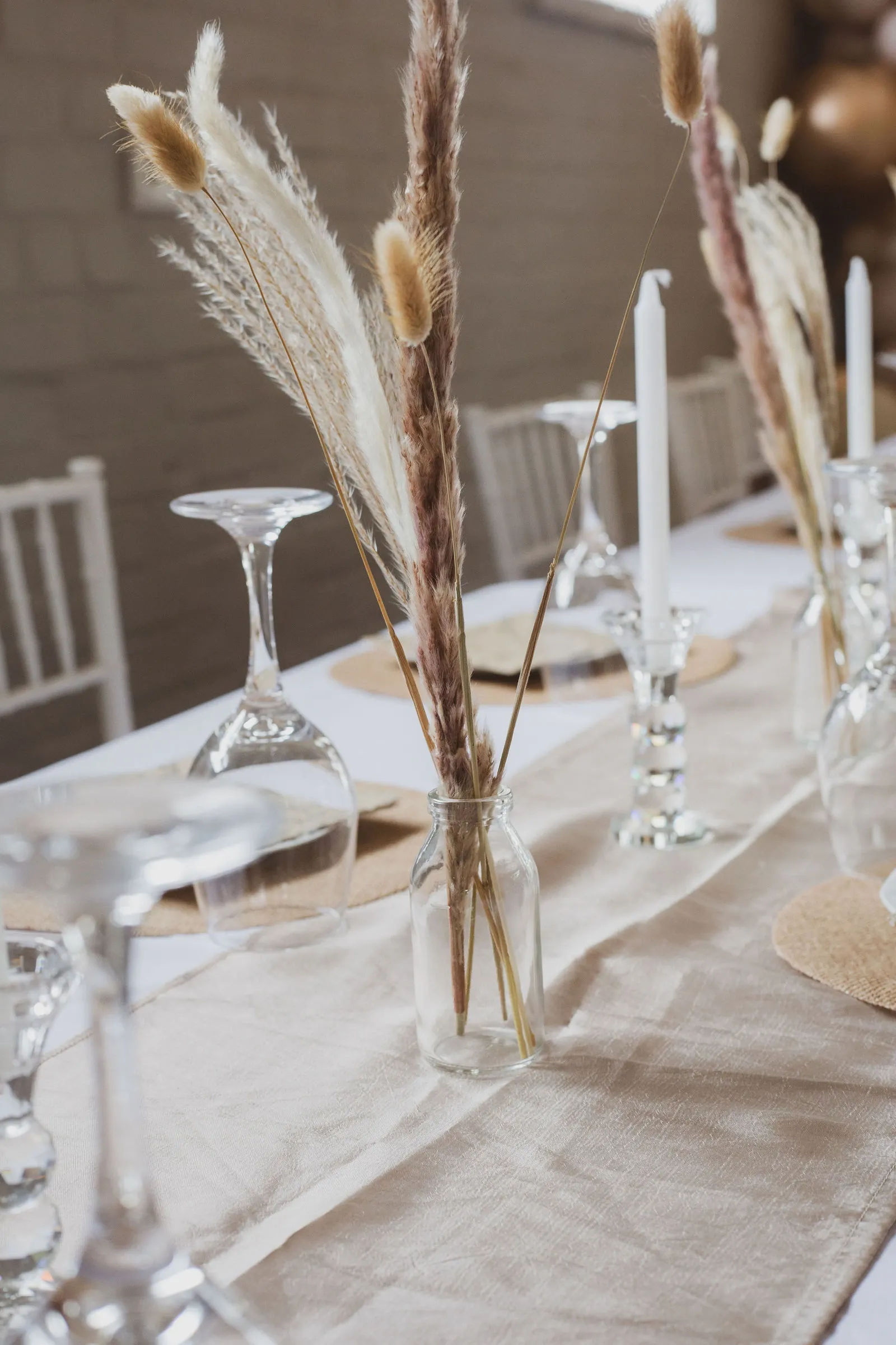 Table detail: dried pampas plumes and bunny-tail grass in glass bottles beside white taper candles, upturned wine glasses and jute place mats.