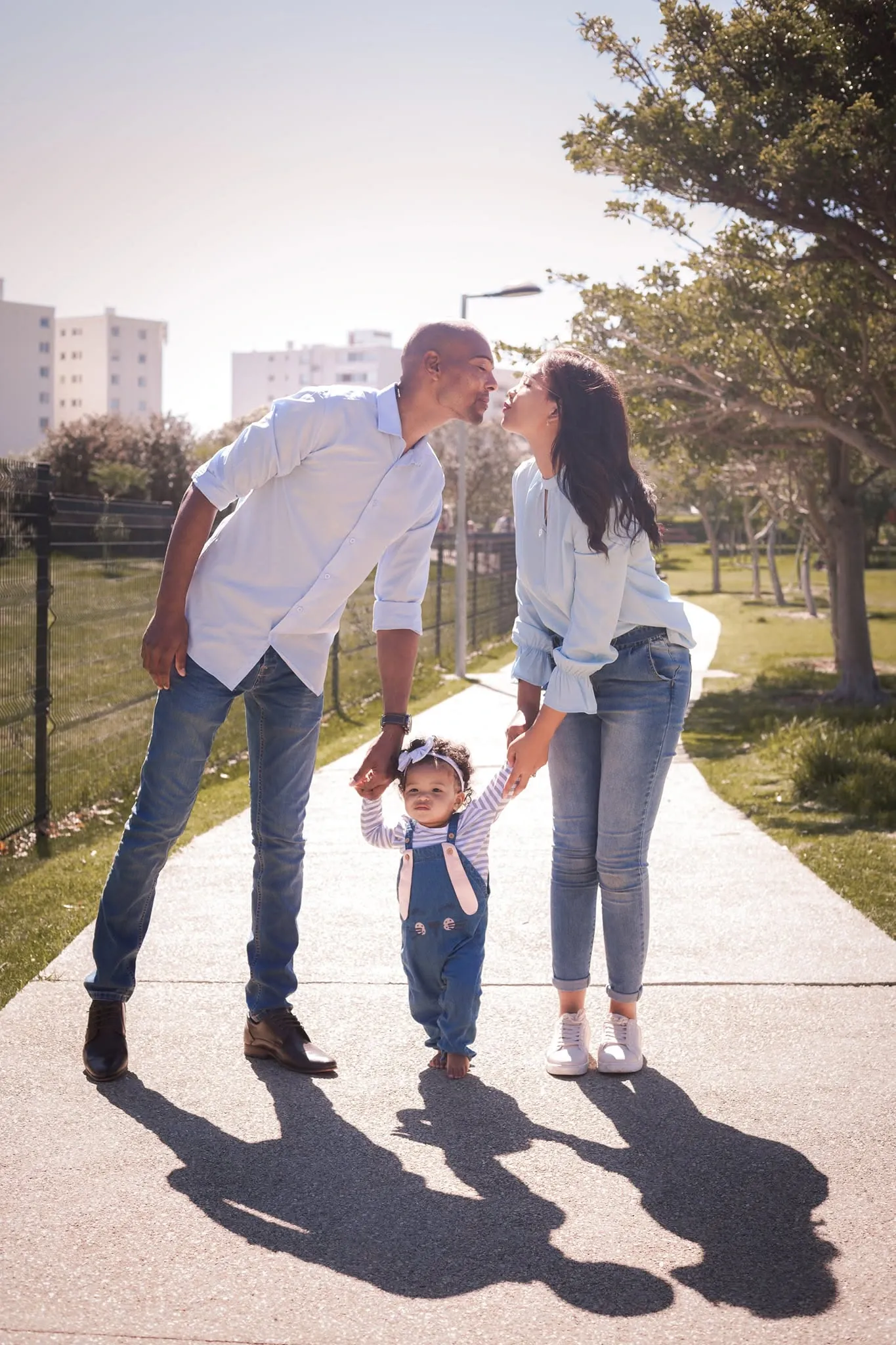 Parents leaning in to kiss over their toddler's head as they walk her by the hands along a sunny path, long shadows on the paving.