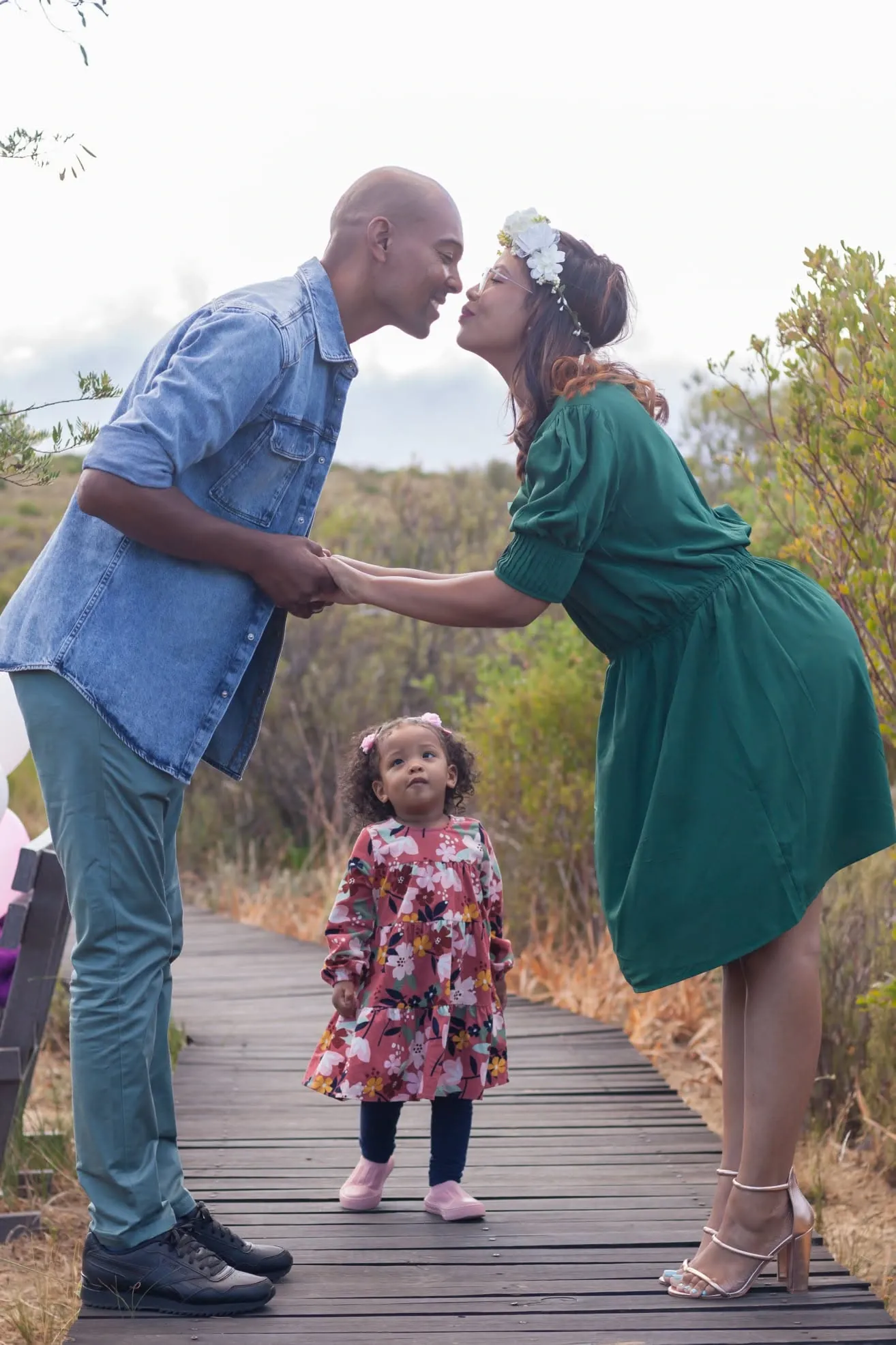 Couple leaning in to kiss on a wooden boardwalk while their toddler daughter in a floral dress looks up at them.