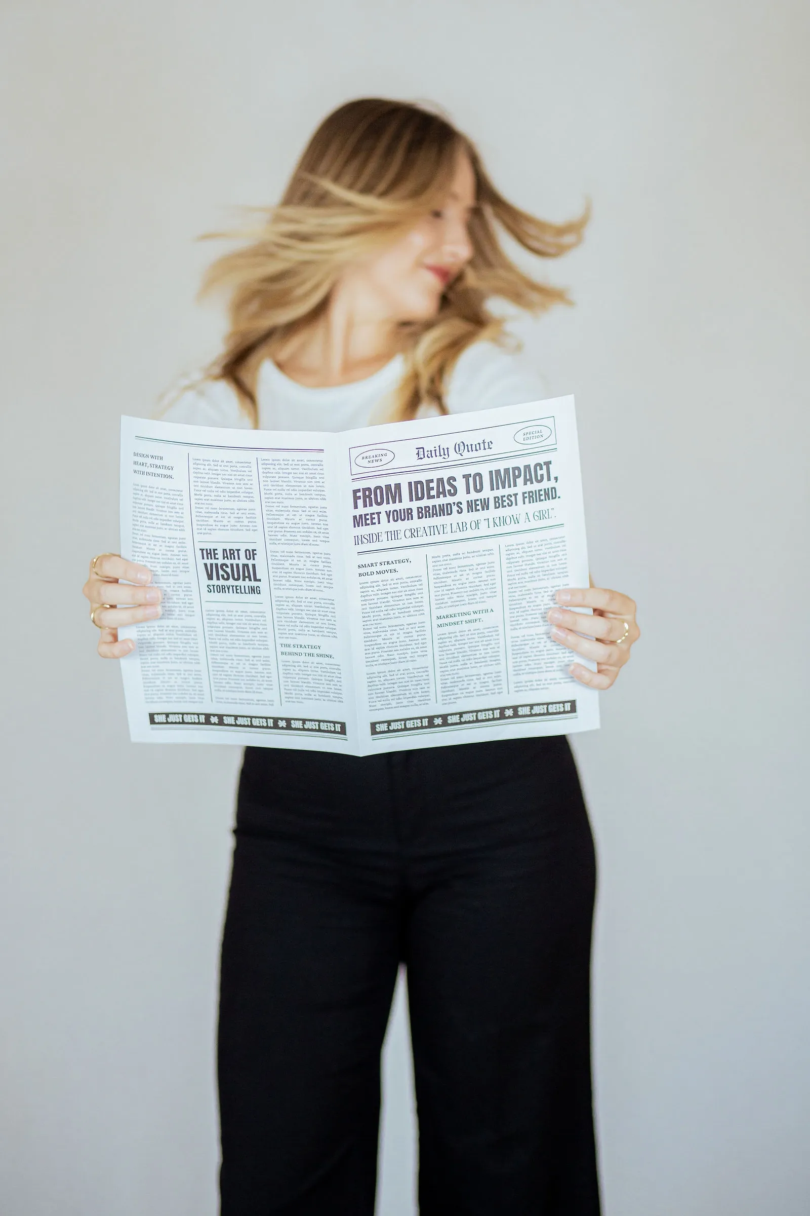 Personal branding photo: a blonde woman flicking her hair while holding an open mock newspaper with marketing headlines.