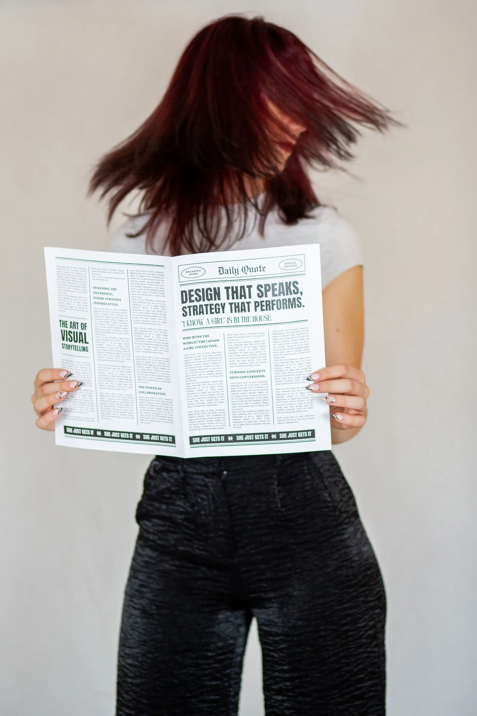 Woman with deep red hair flicking it across her face as she holds an open mock newspaper against a pale backdrop.