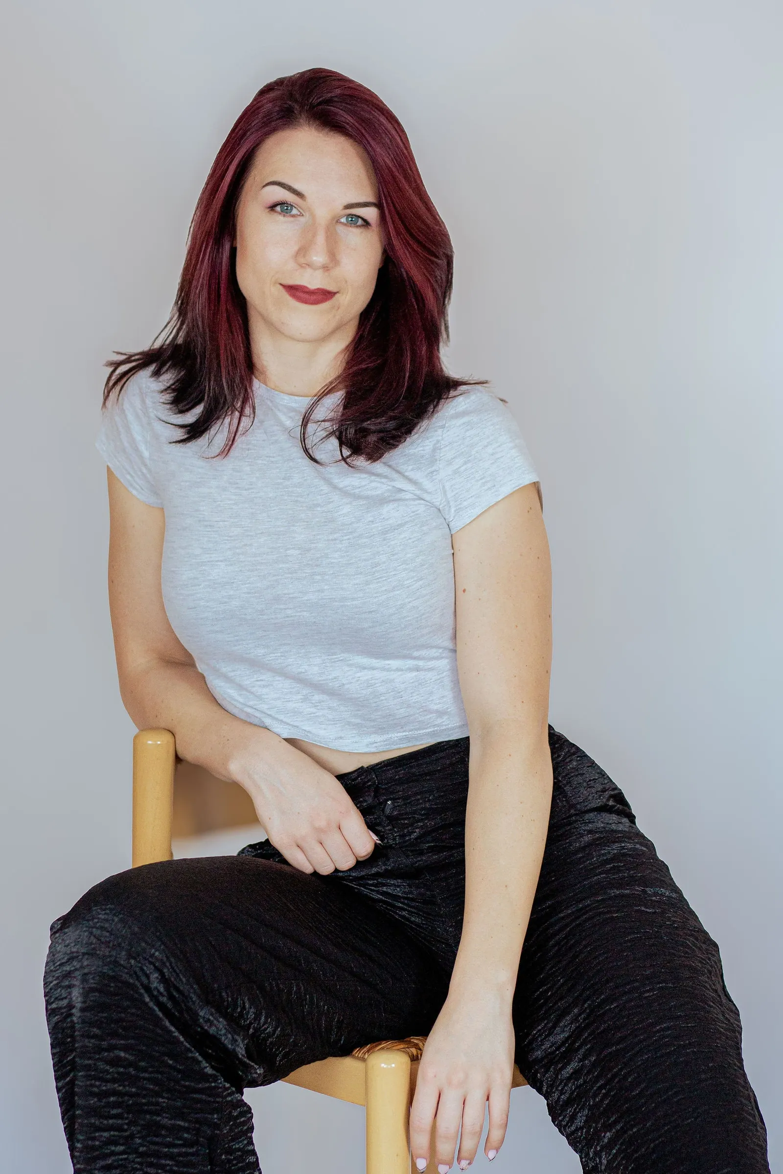 Woman with deep red hair in a grey T-shirt leaning forward on a wooden stool, looking straight at the camera.