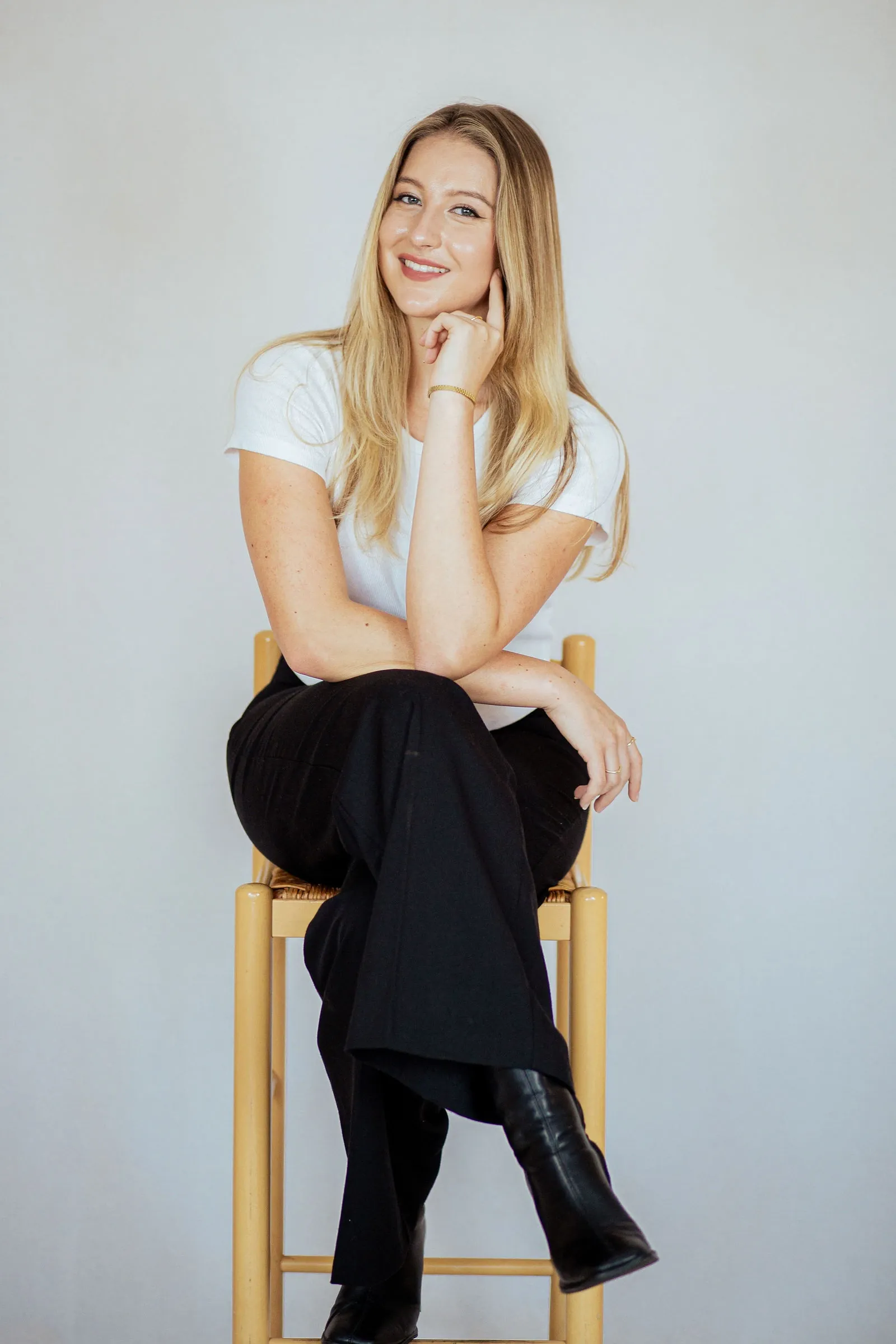 Smiling blonde woman in a white T-shirt seated on a wooden stool, chin on hand and legs crossed.