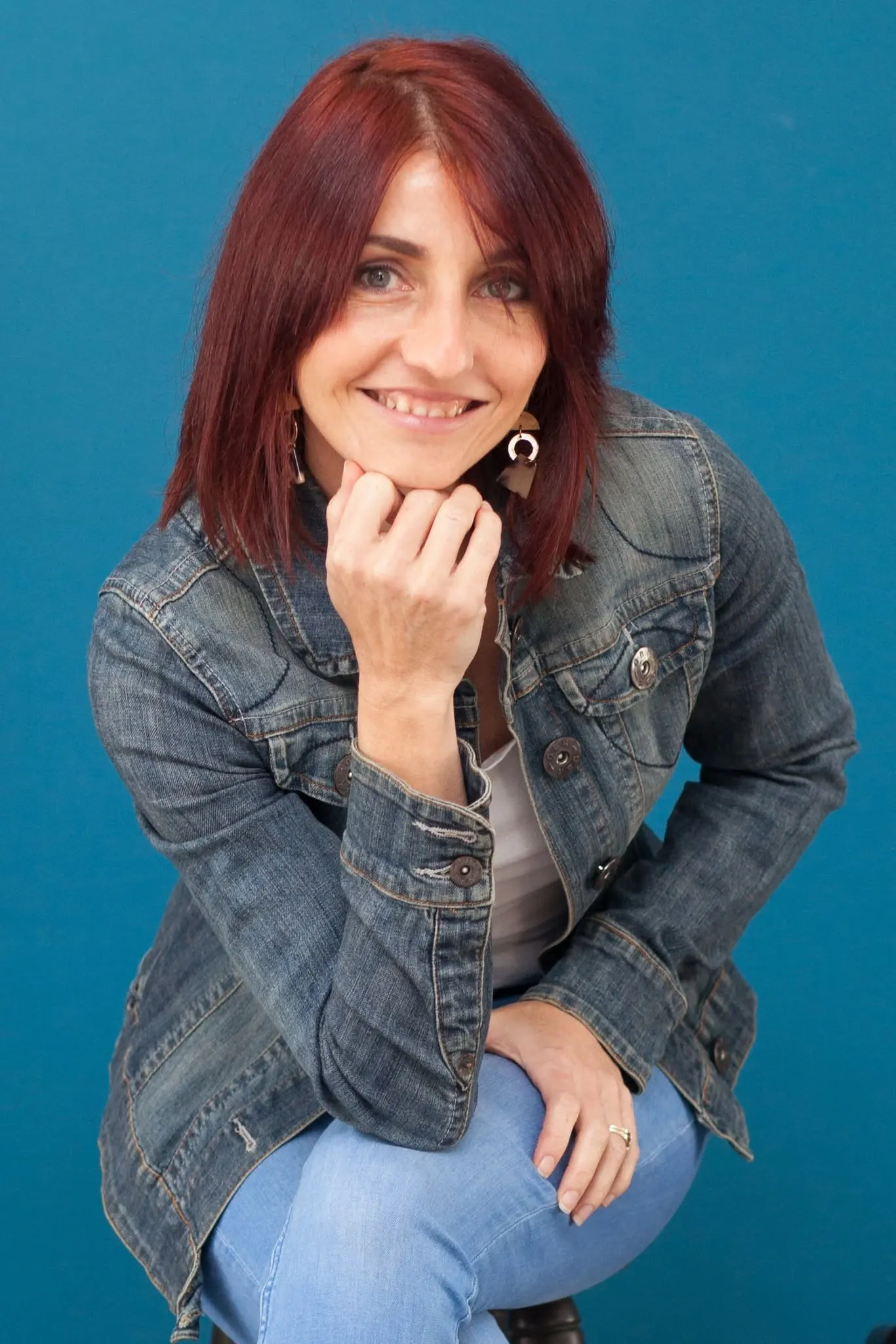 Personal branding portrait: woman in a denim jacket smiling, chin on hand, against a teal wall.