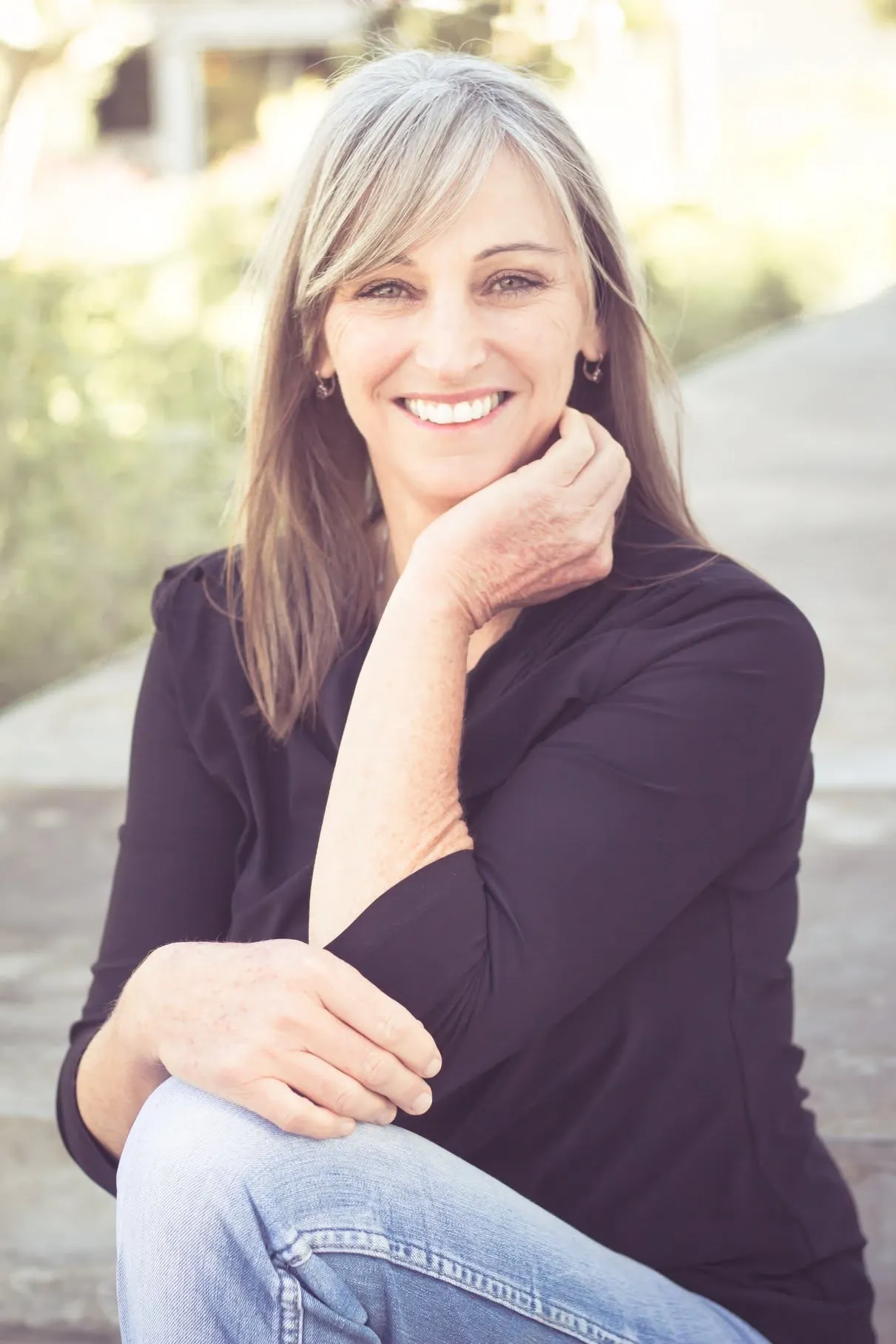Personal branding portrait of a smiling woman with long blonde hair, in a dark blouse and jeans, seated on outdoor steps with her chin resting on one hand.
