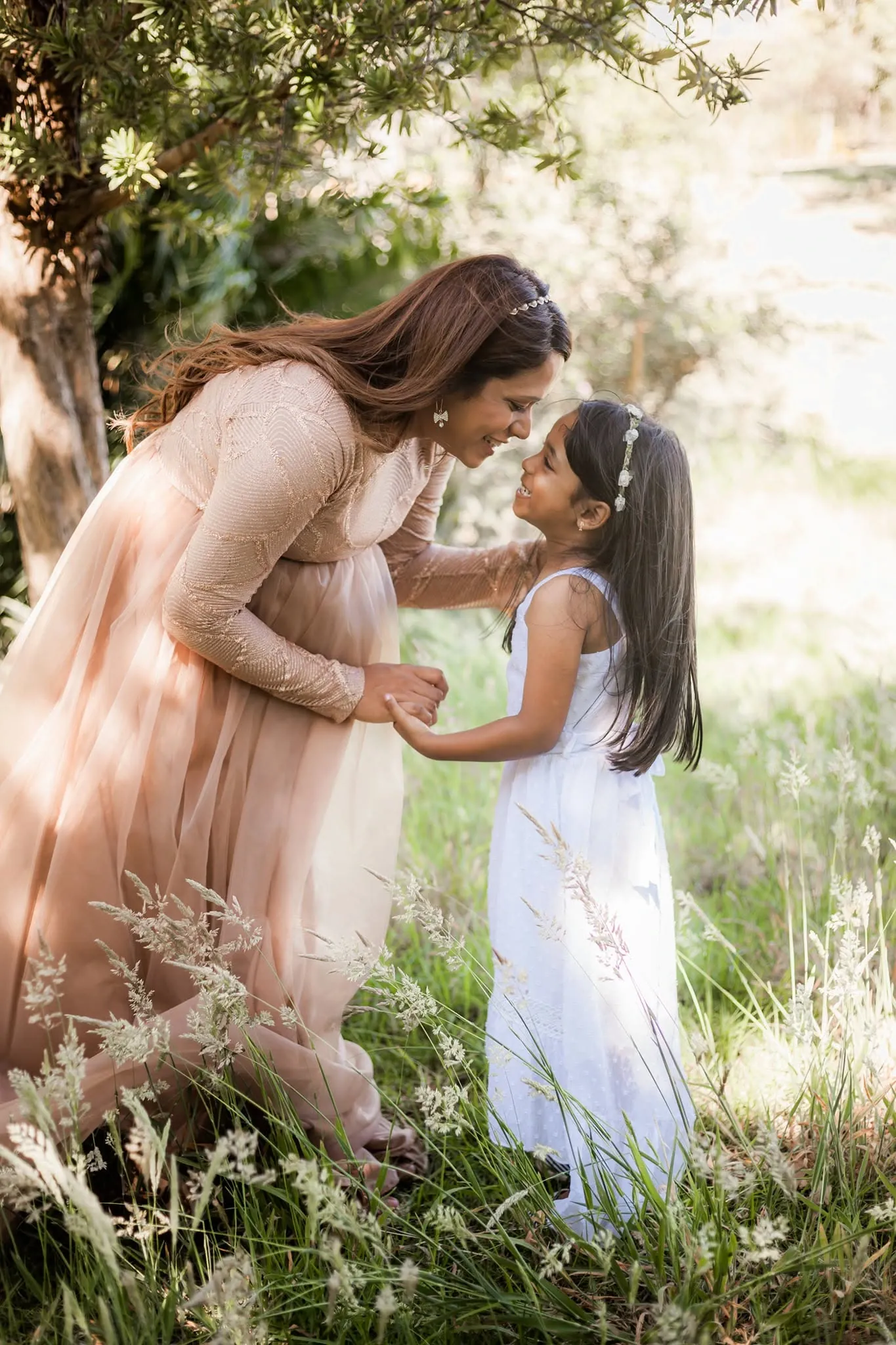 Pregnant mum in a blush gown leaning down nose to nose with her daughter in long grass.