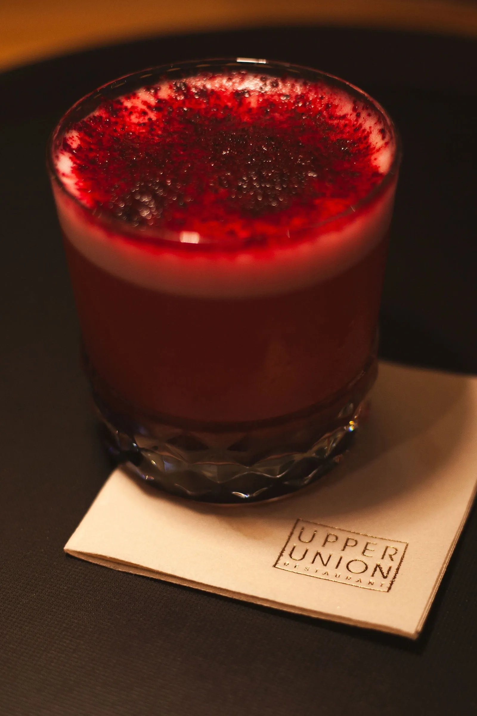 Drinks photography: a deep red cocktail with a pale foam head dusted in red powder, in a cut-crystal tumbler on a printed napkin.