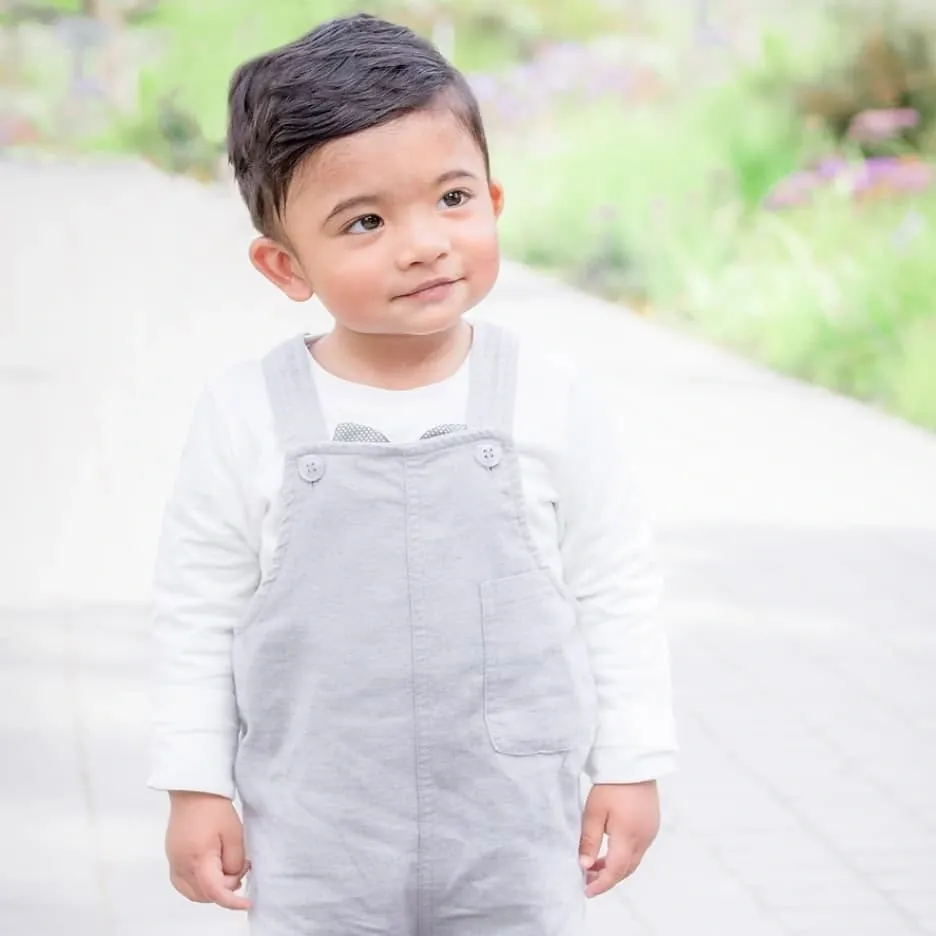 Toddler boy in grey dungarees and a white shirt standing on a path, looking to one side.
