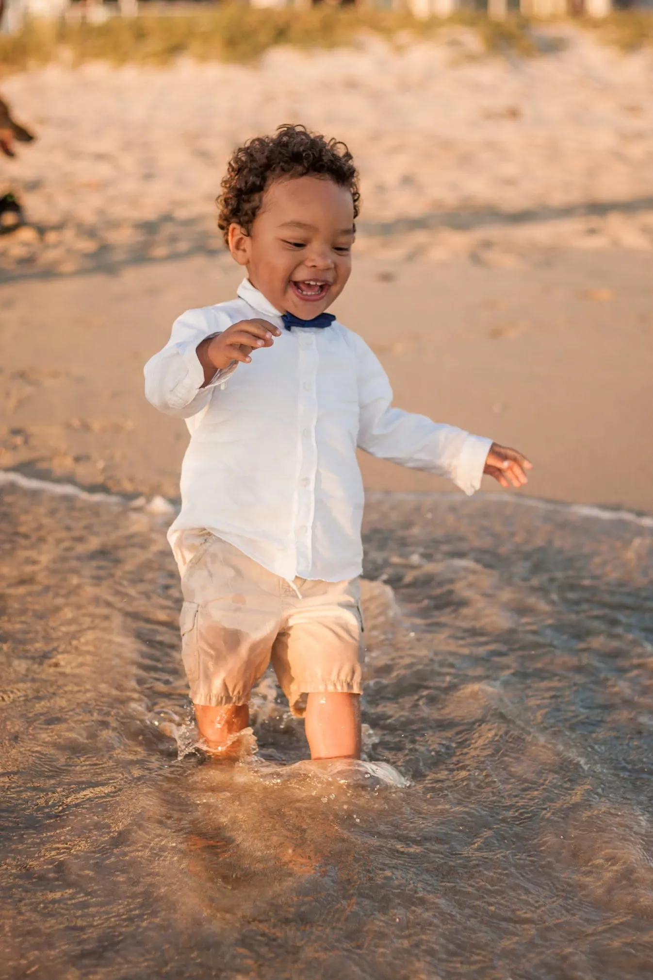 Toddler in a white shirt and bow tie splashing through the shallows at golden hour.
