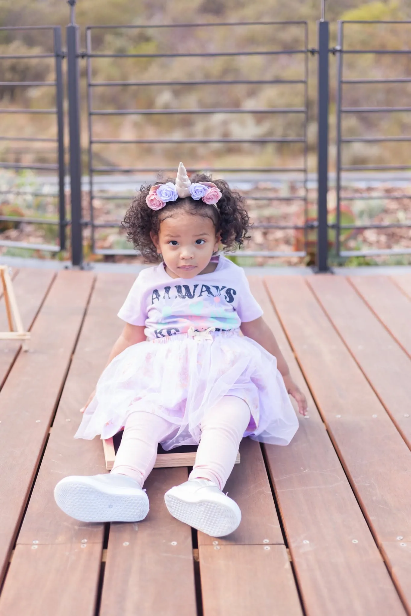 Toddler in a unicorn headband and lilac tutu sitting on a wooden deck.