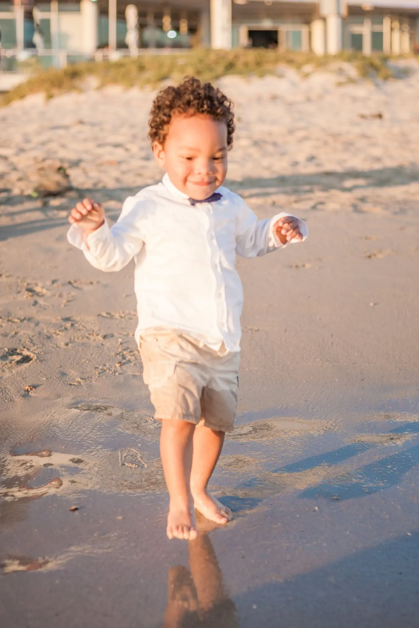Toddler in a white shirt and bow tie walking barefoot across wet sand in warm evening light.